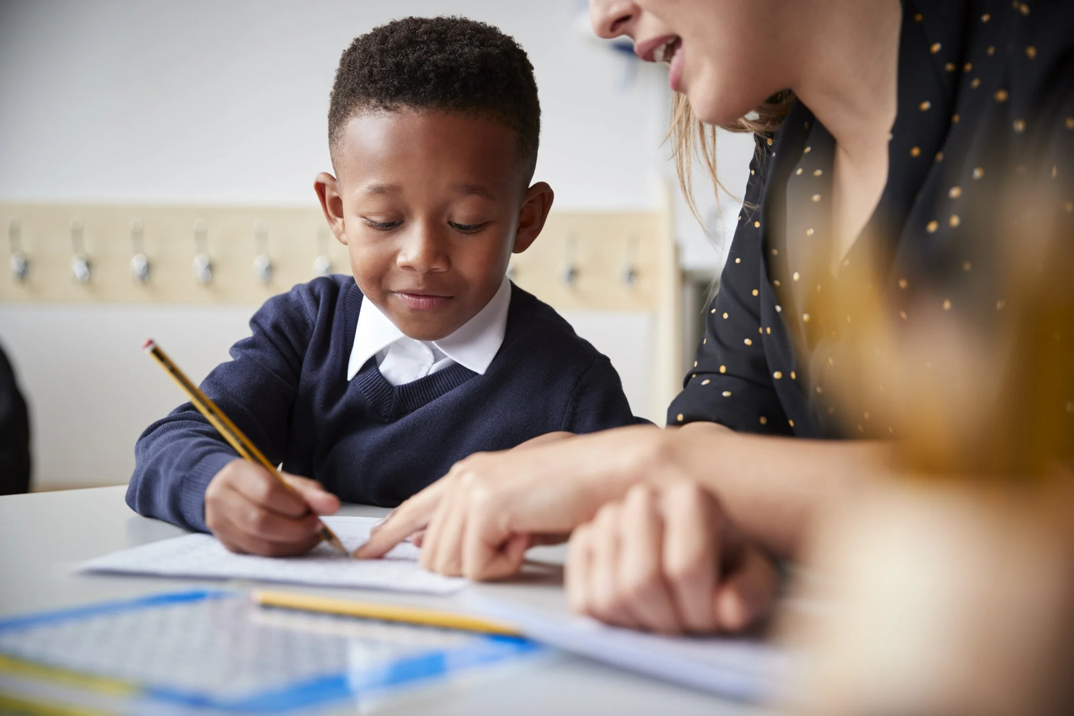 A young boy in a school uniform writing with a pencil, with an adult woman assisting him, at a desk in a classroom.