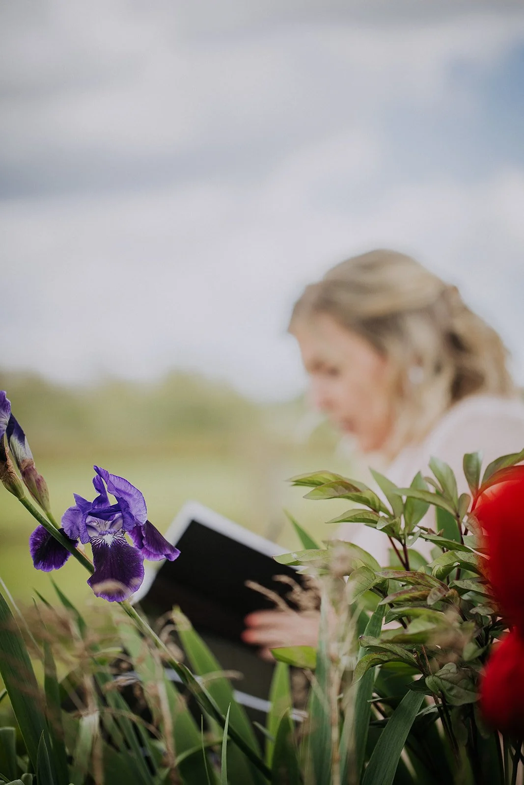 A woman with blonde hair reading a black book or notebook outdoors among purple flowers and green foliage, with a cloudy sky in the background.