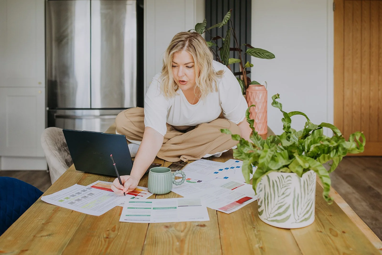 Woman sitting on a wooden table, working with documents, a laptop, and a calculator, with a large potted plant and a pink vase nearby in a kitchen or dining area.