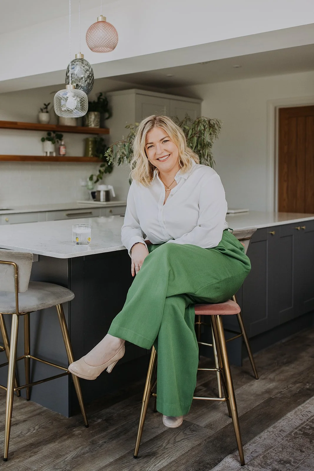 Laura Cresswell, founder of The Lume Collective, sitting on a stool in a kitchen and smiling at the camera.