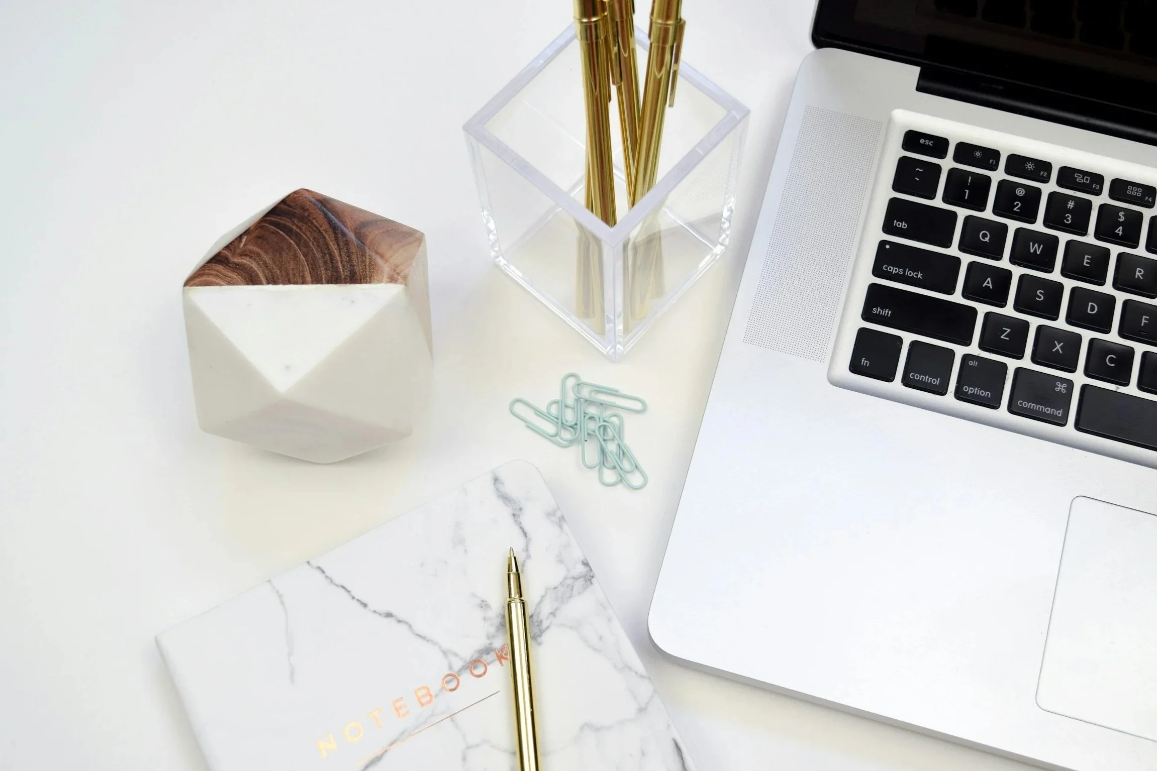Flat lay of a laptop, notebook, pens, and paper clips on a white desk.