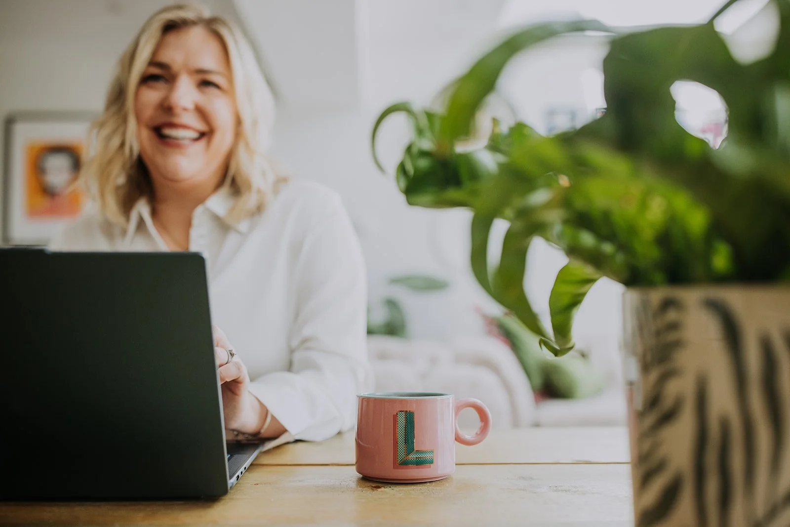 Laura Cresswell smiling at a laptop at a desk, with a houseplant in the foreground.