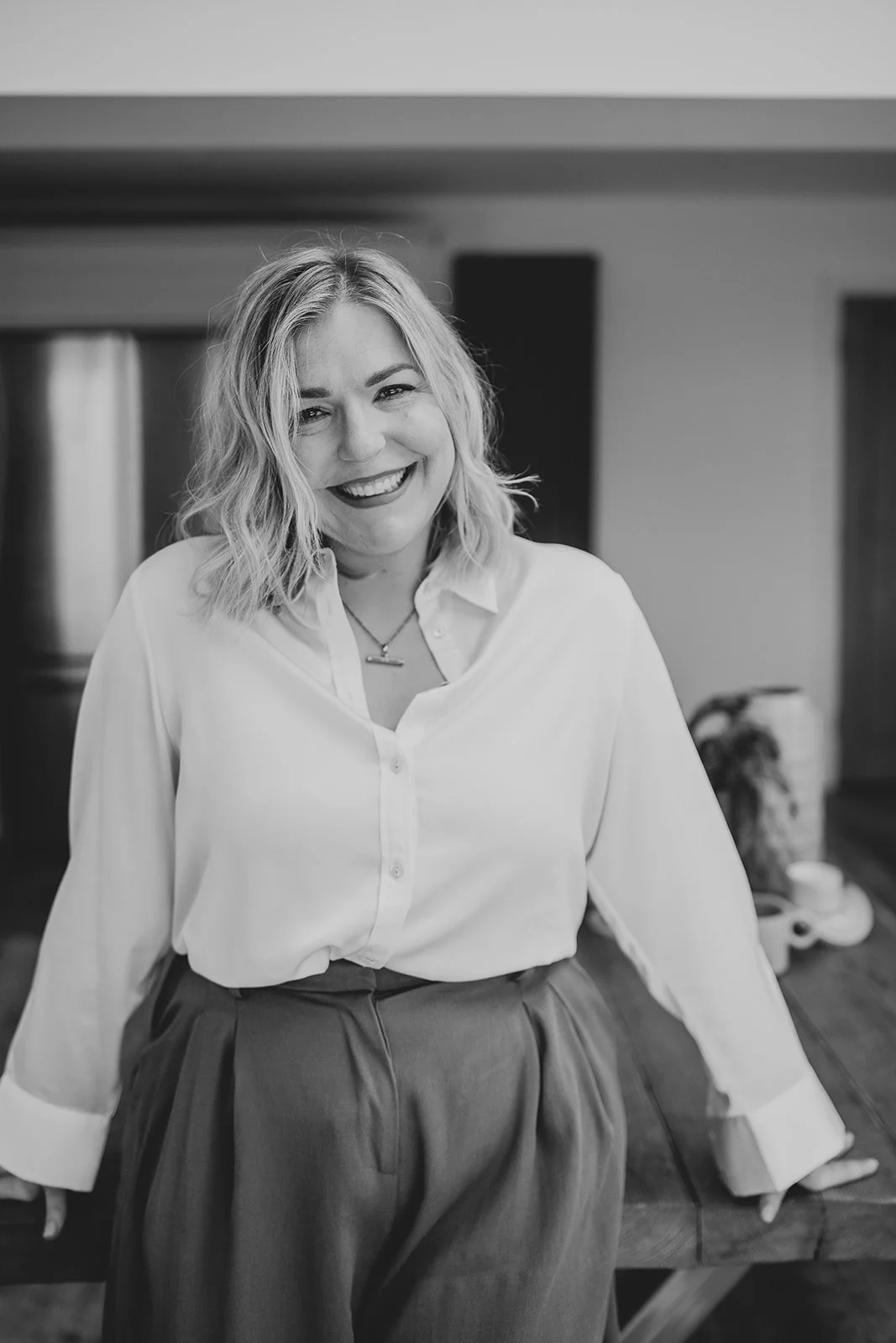 A woman with shoulder-length wavy hair smiling while standing indoors, wearing a white button-up shirt and pleated pants.