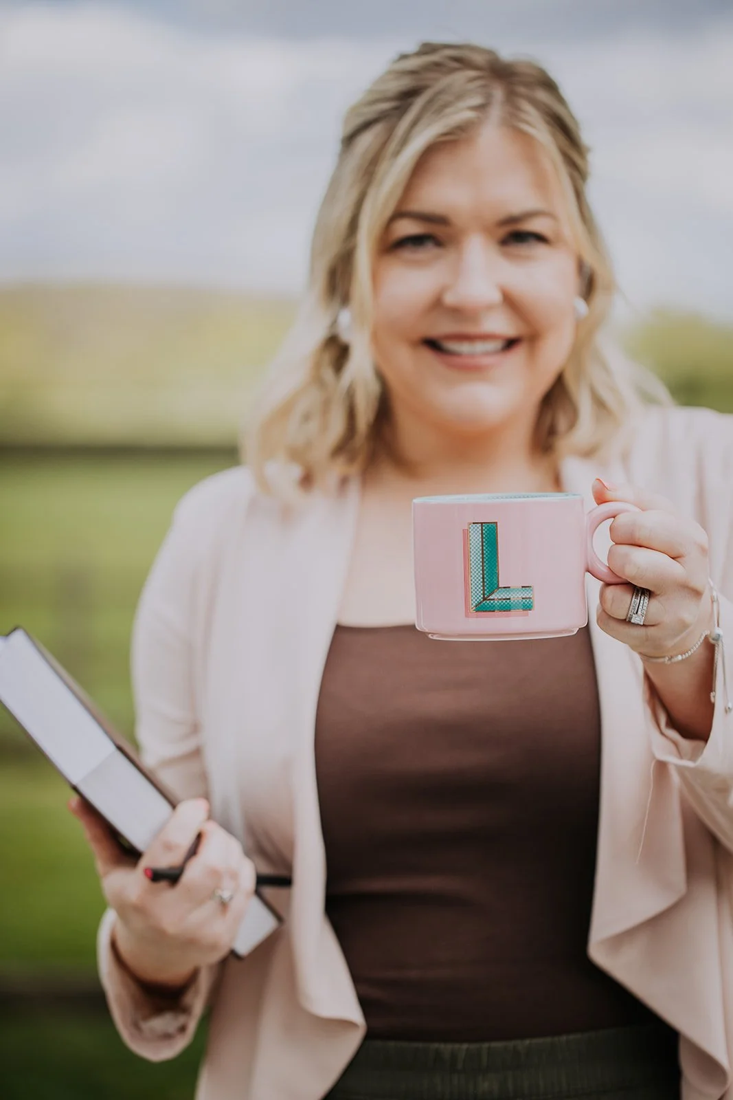 A woman holding a pink mug with a blue letter 'L' on it and a notebook in her other hand, outdoors with greenery and a cloudy sky in the background.