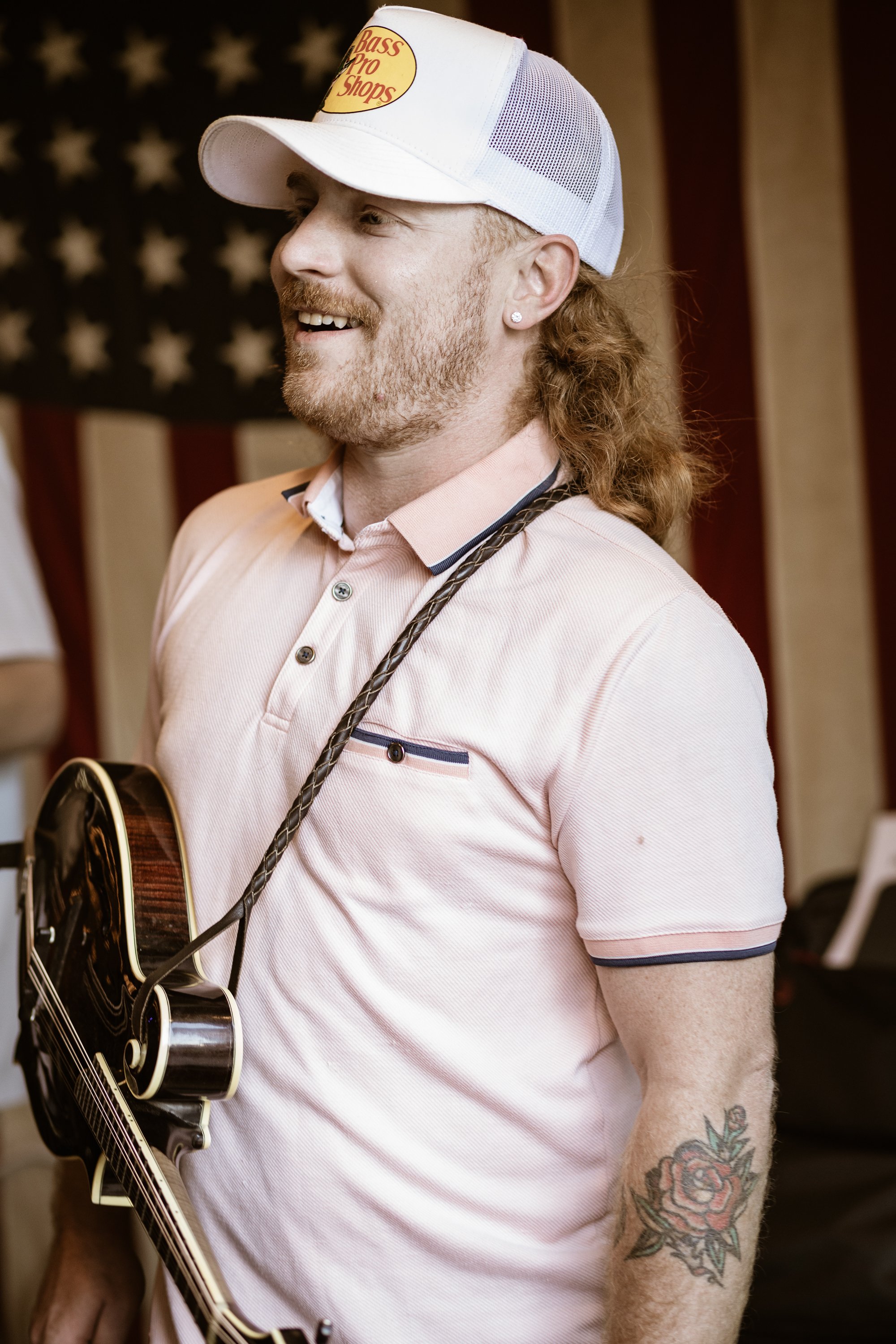 Tristen Norfleet, with long, curly red hair wearing a white cap with a Bass Pro Shops logo, smiling, holding an acoustic guitar, and standing in front of an American flag.