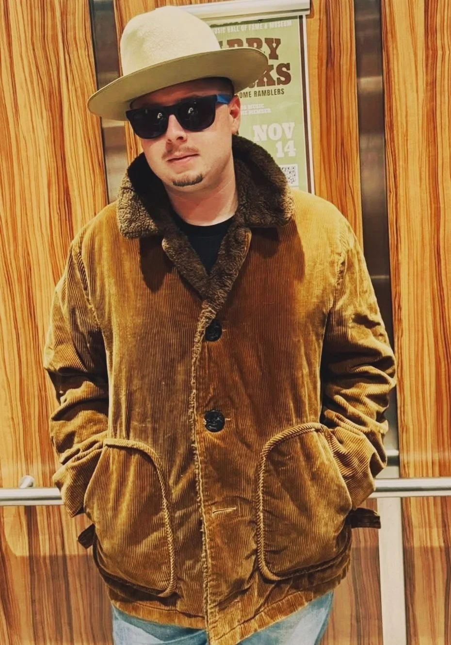 Kelsey Crews, wearing a beige wide-brimmed hat, black sunglasses, a brown corduroy jacket with a fur collar, and light blue jeans, is standing in an elevator with wood paneling.
