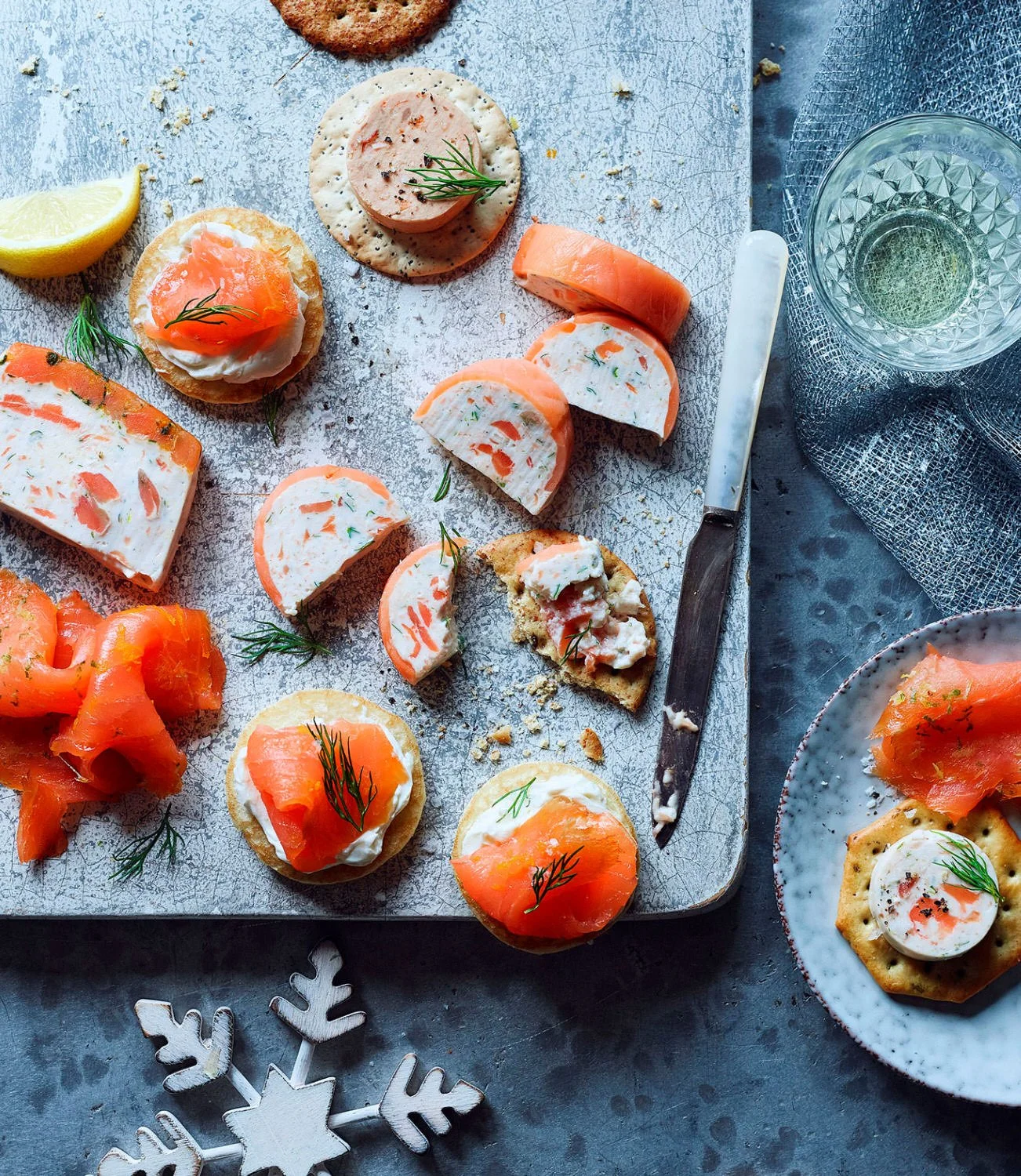 Assorted smoked salmon and cream cheese appetizers on a gray surface, garnished with herbs, with a lemon wedge, a glass of water, and a decorative snowflake ornament nearby.