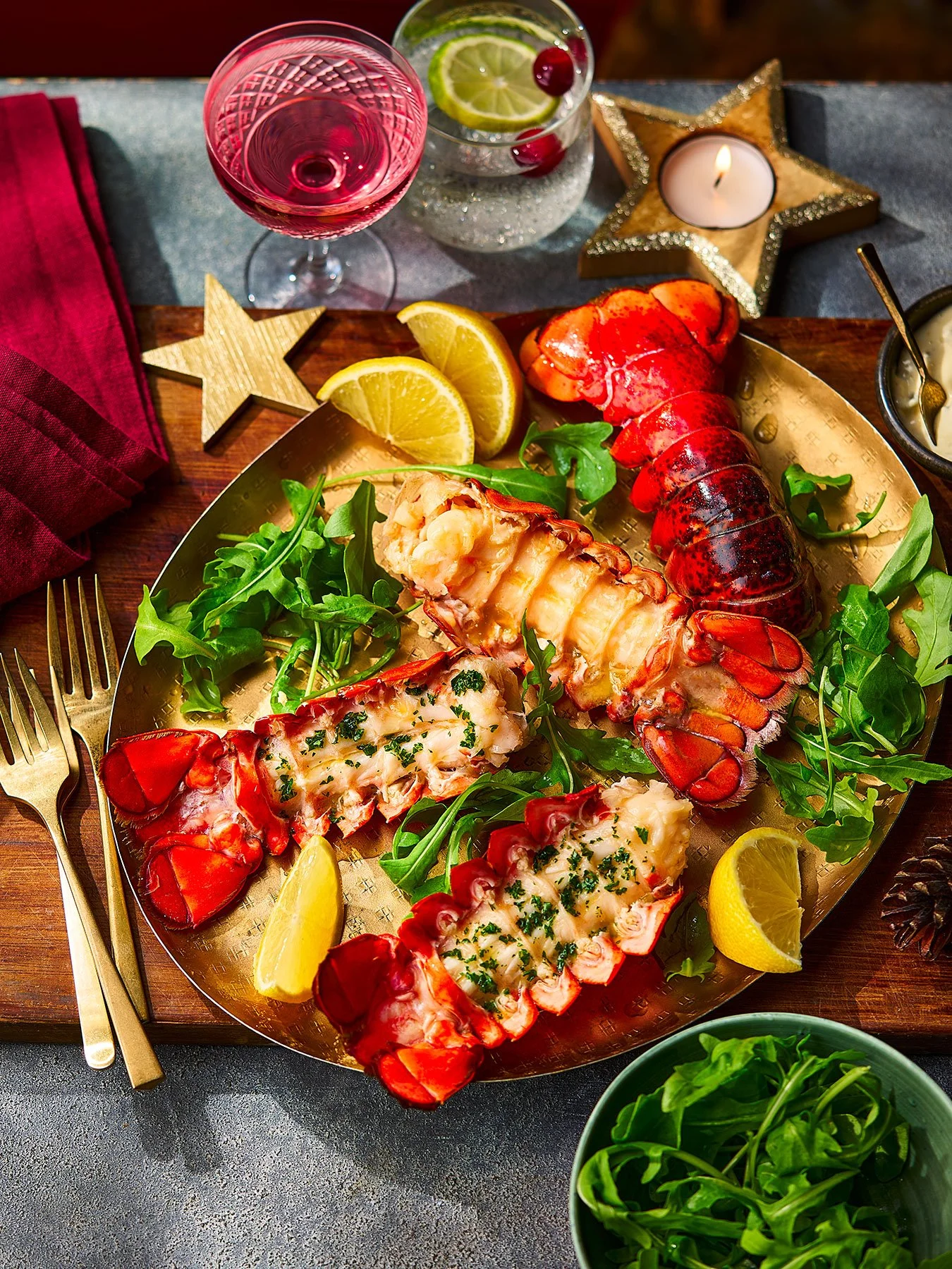 Lobster tails garnished with herbs, served on a golden platter with lemon wedges, arugula, and glasses of wine and a cocktail, festive table setting with candle and star decorations.