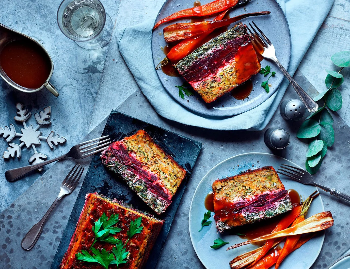 Slices of meatloaf with sauces and roasted vegetables on plates and a serving dish on a table with utensils, salt, and decorative elements.