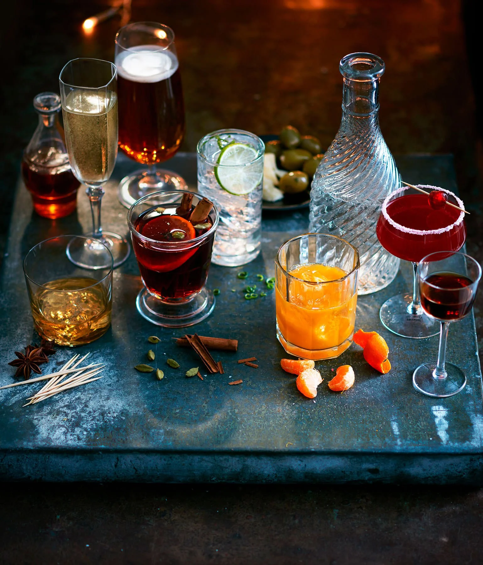 A variety of colorful cocktails and drinks on a dark table, including a yellow-orange beverage, a red drink with a cherry garnish, a clear drink with lime slices, and others, along with a decanter, olives, and spices.