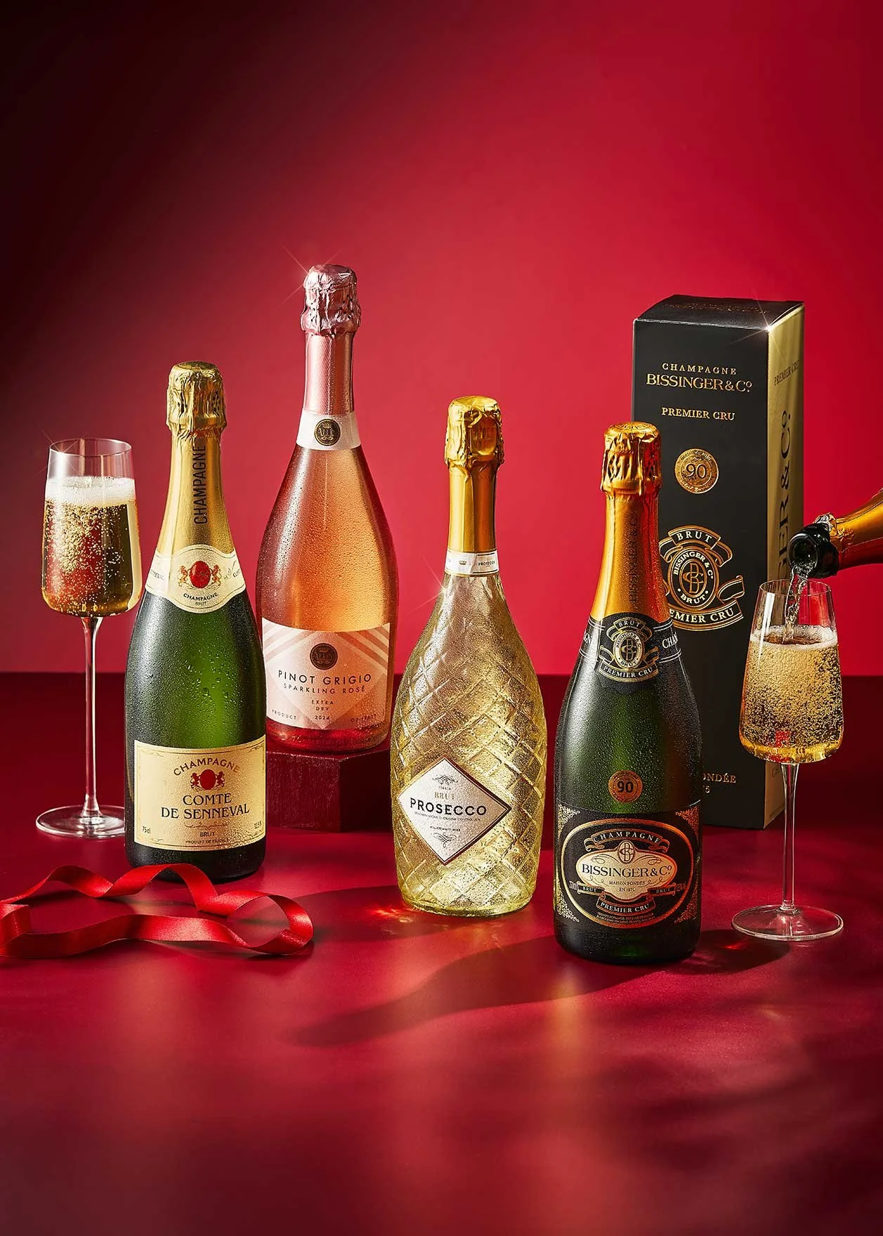 Various bottles of champagne and sparkling wine with two glasses filled with bubbly, set against a red background.