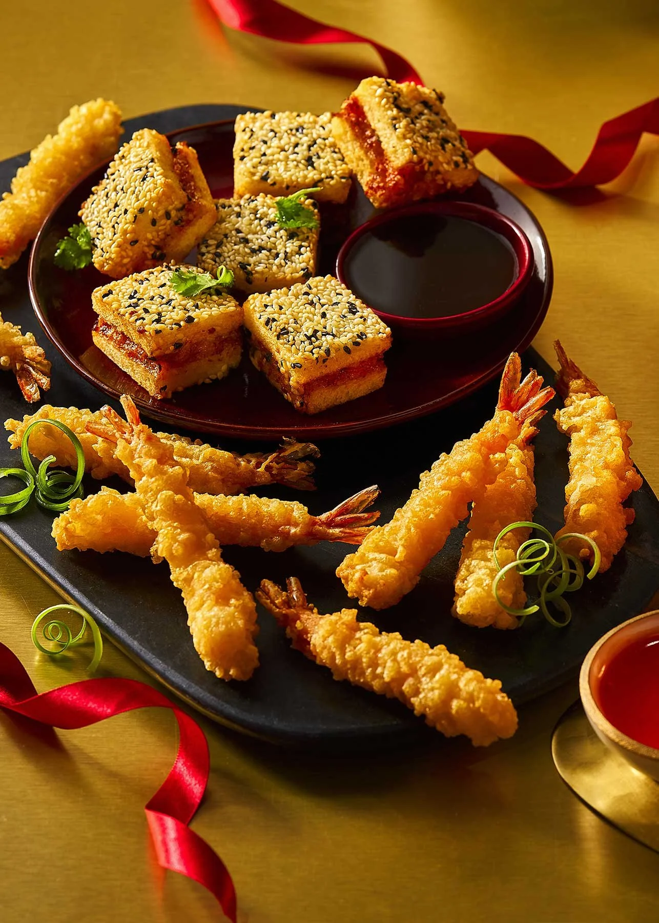 Fried shrimp tempura and sesame-crusted cheesecake bites served with soy sauce and dipping sauces on black and red plates, decorated with green garnishes and red ribbons.