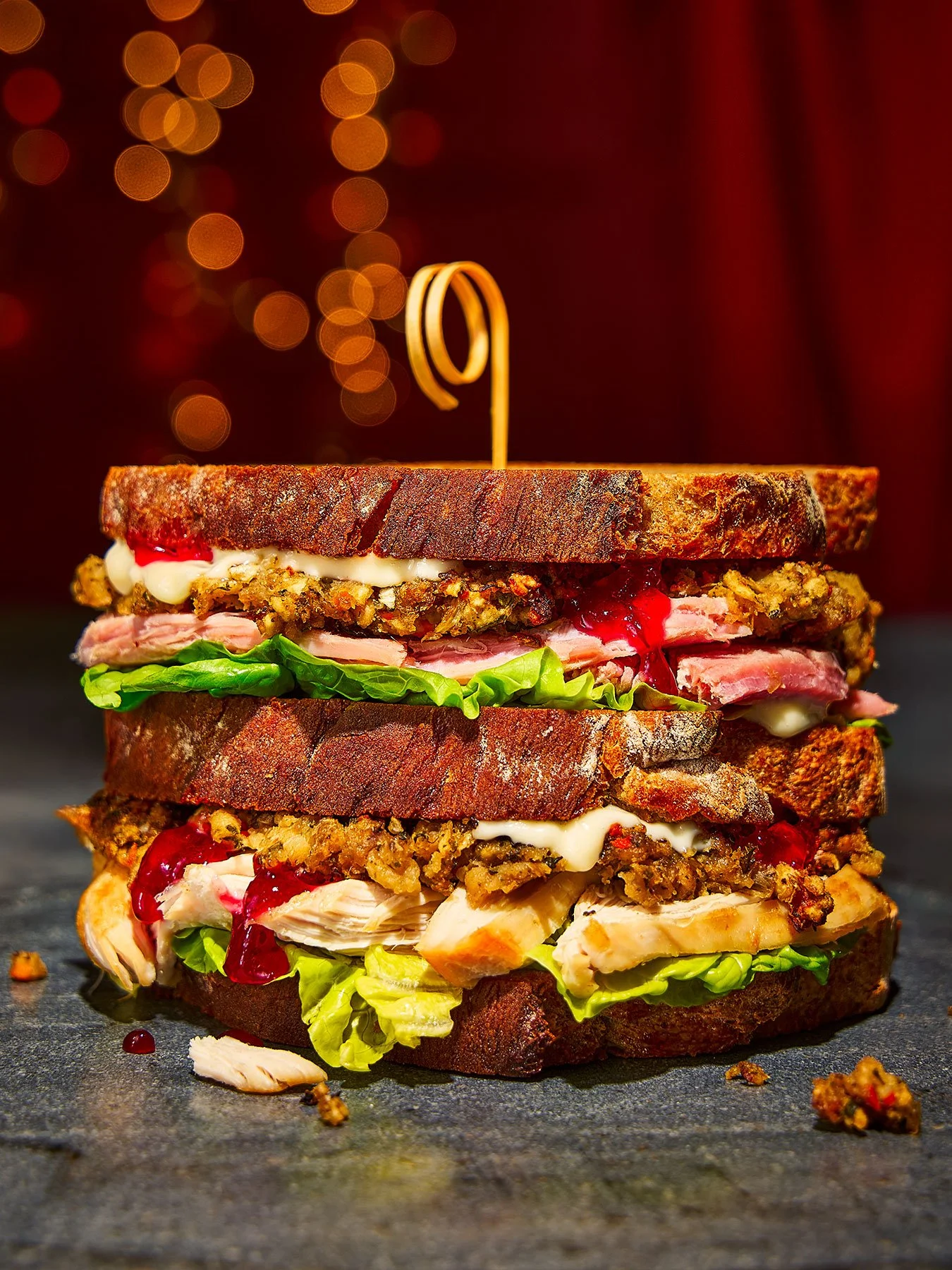 A towering multi-layered burger with various meats, lettuce, and condiments, topped with a pretzel bun and a decorative skewer, on a dark surface with bokeh lights in the background.