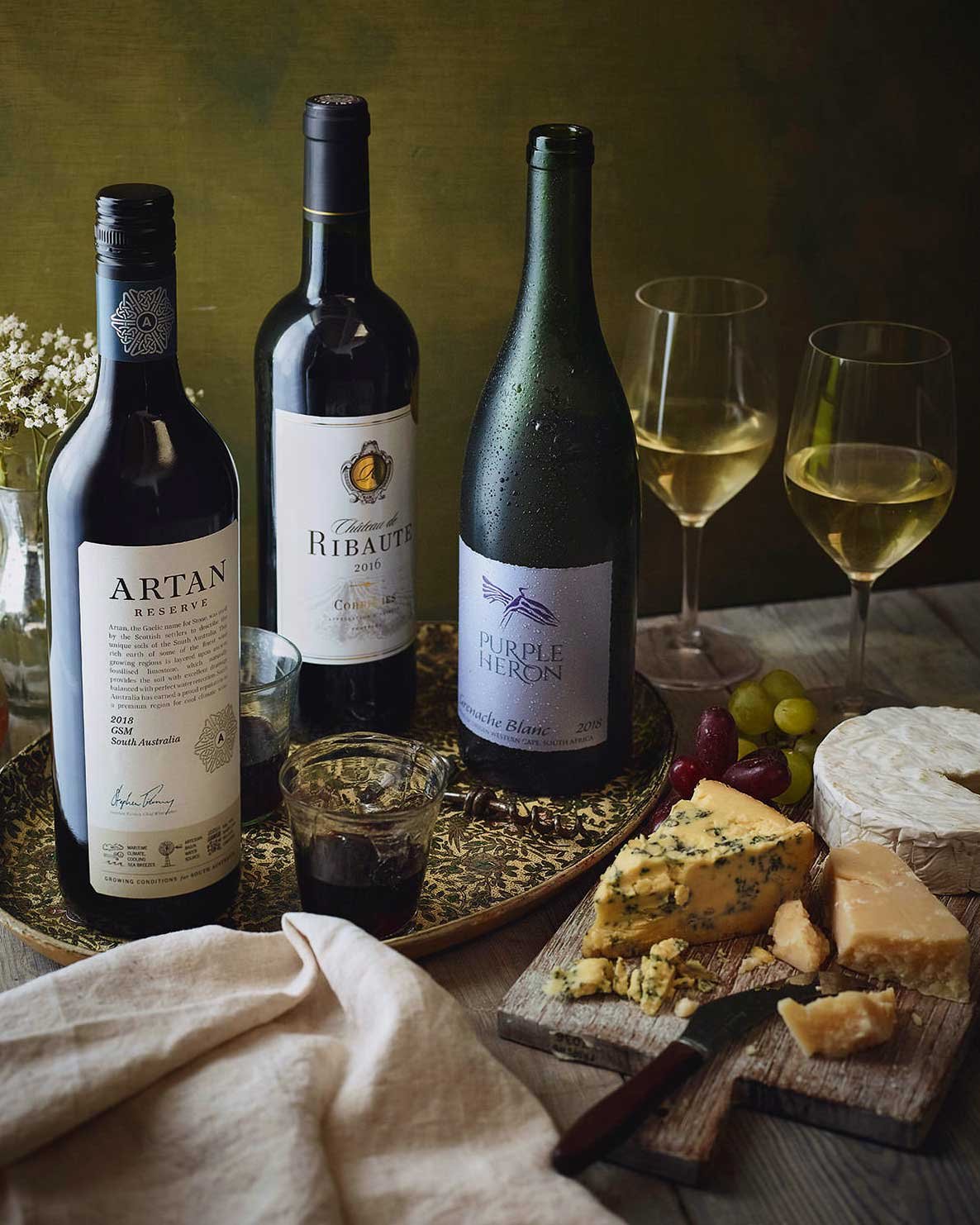 Three bottles of wine, two glasses of white wine, assorted cheeses, grapes, and a small bouquet of flowers on a wooden table.
