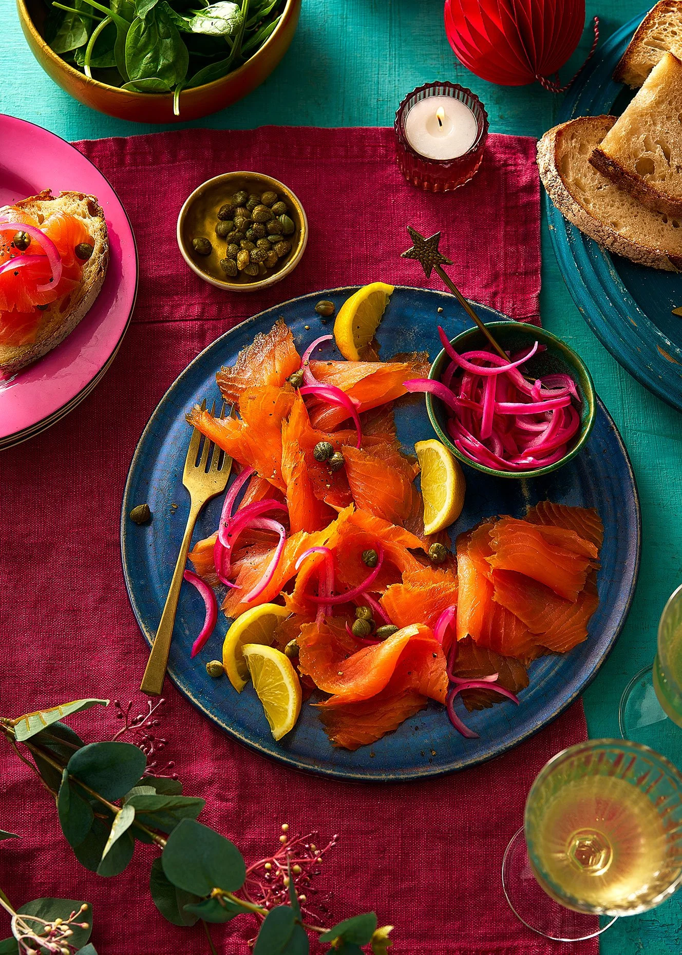 A table setting with smoked salmon slices, lemon wedges, pickled onions, capers, a small bowl of green olives, a candle, a glass of white wine, and slices of bread on colorful plates and a red and teal tablecloth.