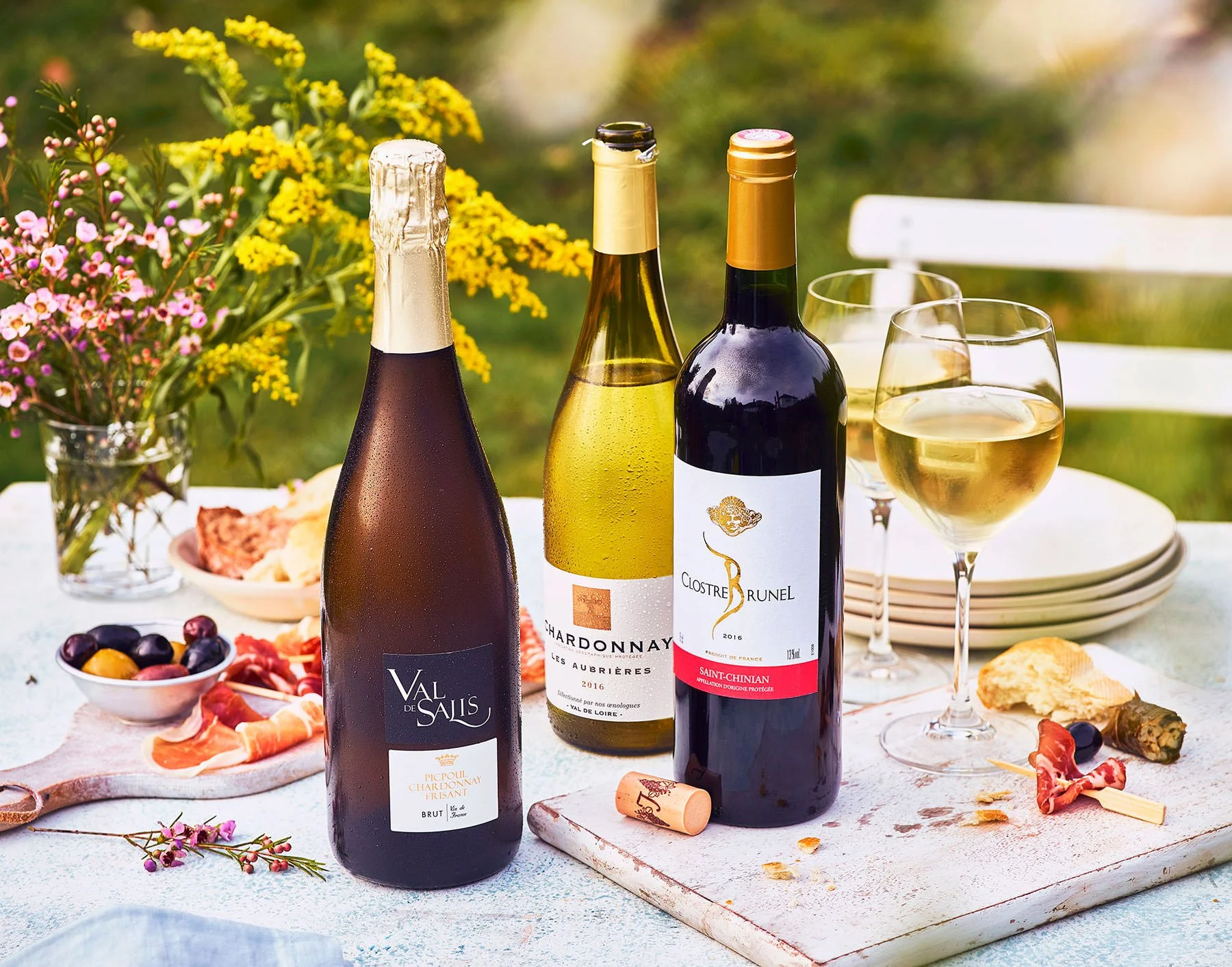 Bottles of sparkling wine, Chardonnay, and red wine on a table outdoors with glasses of white wine, a bowl of grapes, sliced cured meats, and a cheese wedge, surrounded by flowers and plates.