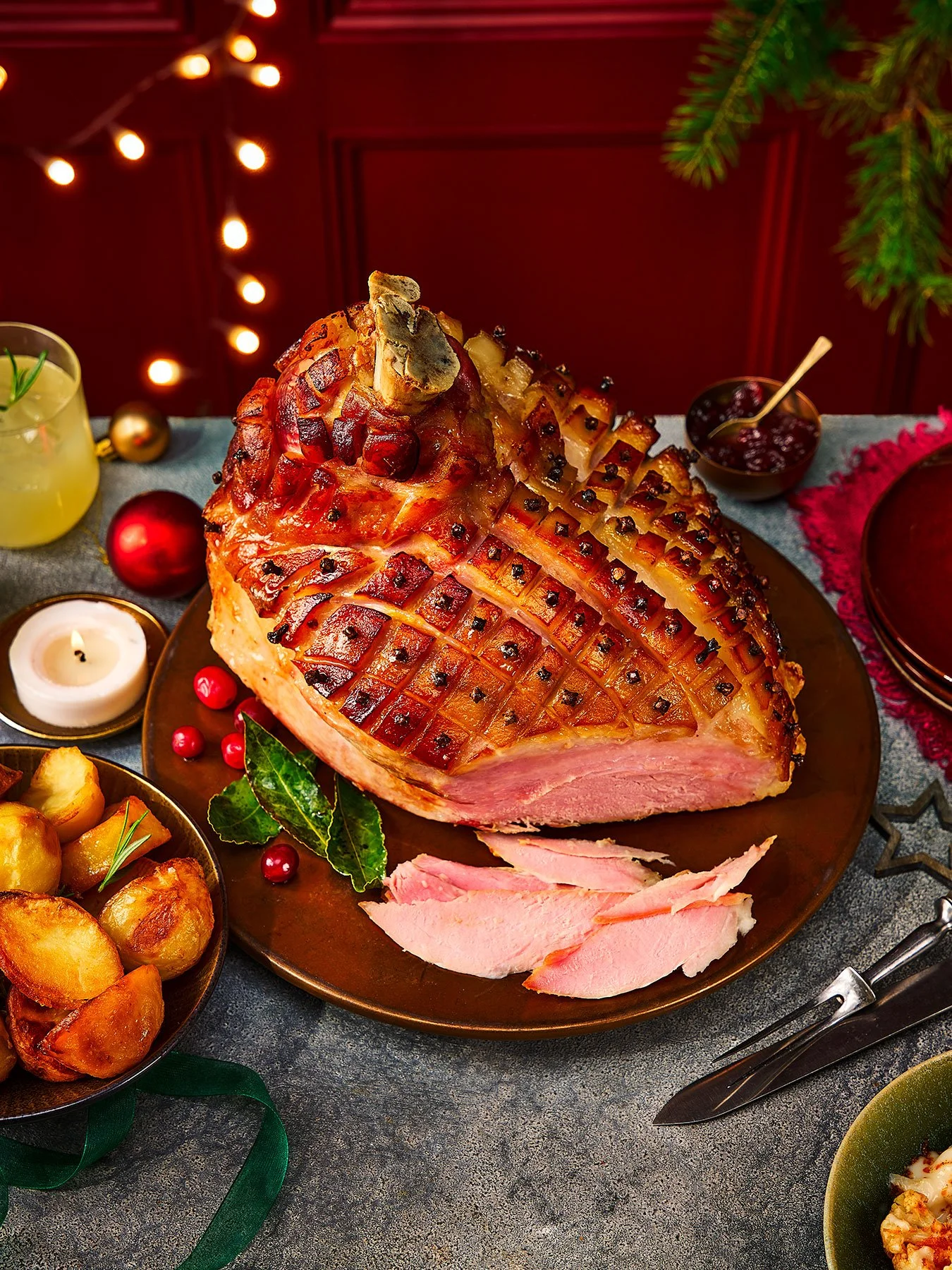 Baked ham with a lattice pattern, garnished with herbs, on a dinner plate with sliced ham beside it, surrounded by Christmas decorations and side dishes.