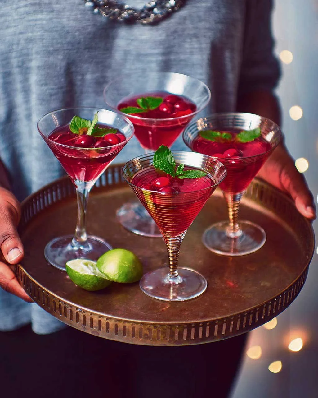 A person holding a round copper tray with four cranberry cocktail drinks garnished with mint leaves and cranberries, and two lime halves on the tray.