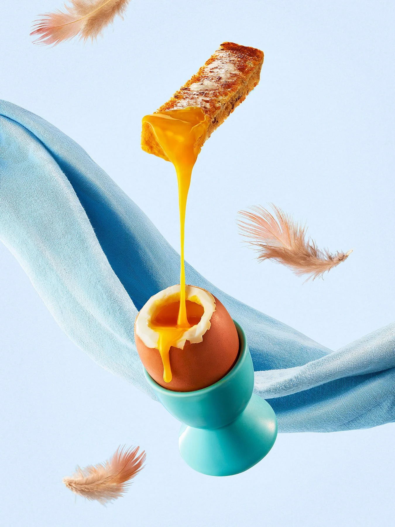 A soft-boiled egg in an egg cup with a cracked shell, with yellow egg yolk flowing into the cup. A piece of fried food, possibly a fish stick or mozzarella stick, is dropping into the yolk, and there are feathers floating around. The background is li