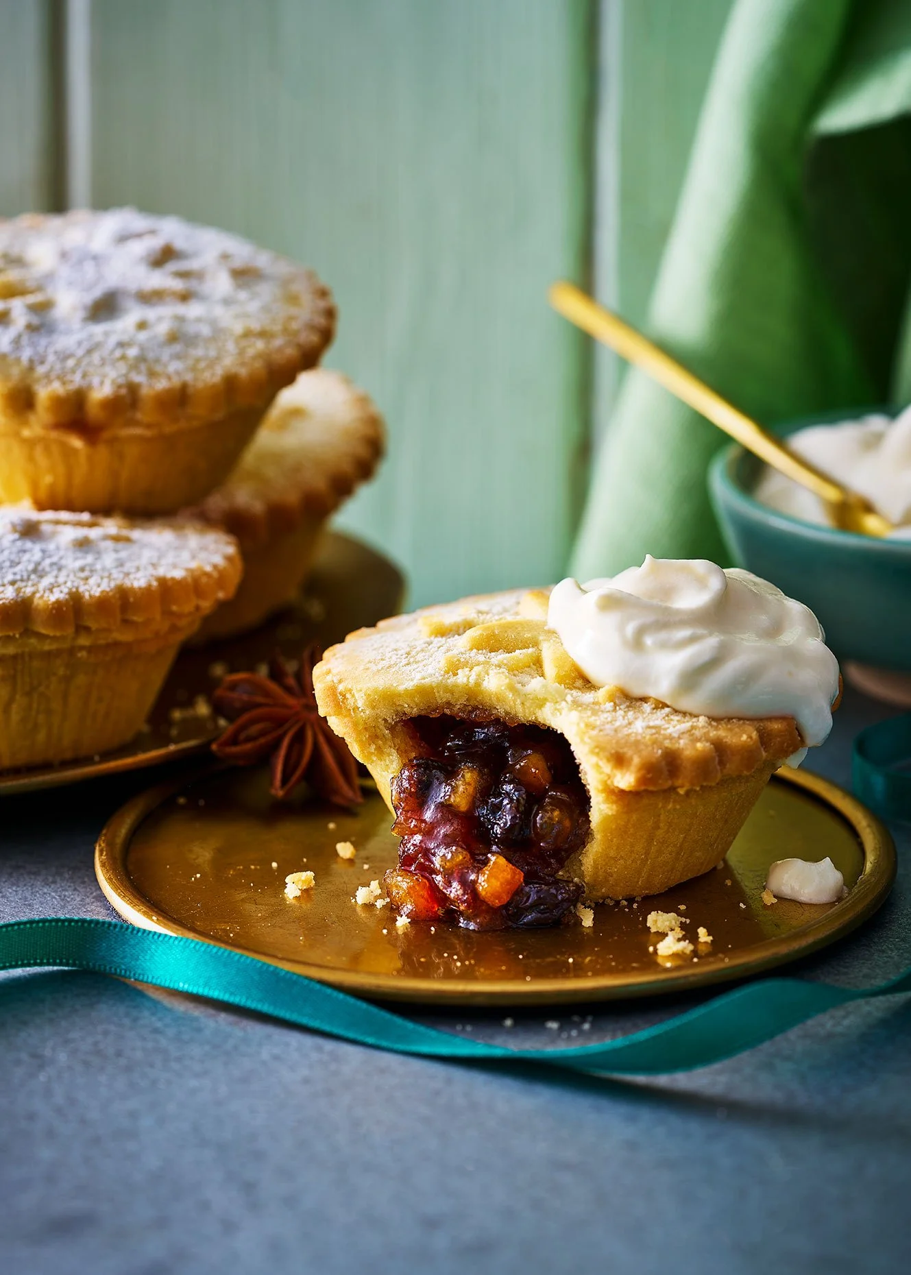 A sliced cherry pie with whipped cream on top, placed on a gold plate with empty pie crusts, star anise, and a teal ribbon.