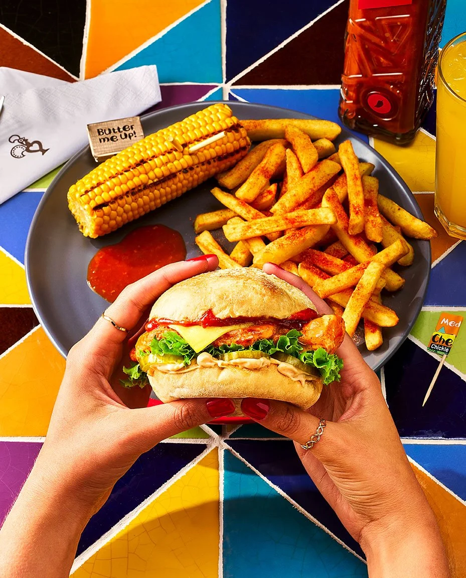Person holding a sandwich with lettuce, tomato, cheese, and chicken. In front of a plate with French fries, a corn on the cob with a 'Butter me up!' sign, and ketchup. Beverages include a red drink and a yellow drink. The background features a colorf