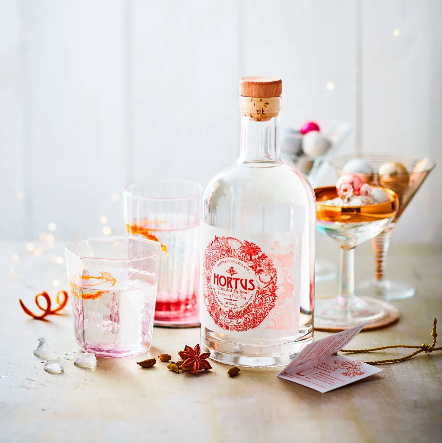 A bottle of Hortus gin surrounded by glasses with pink liquid, star anise, cardamom, and decorative items on a light-colored surface.