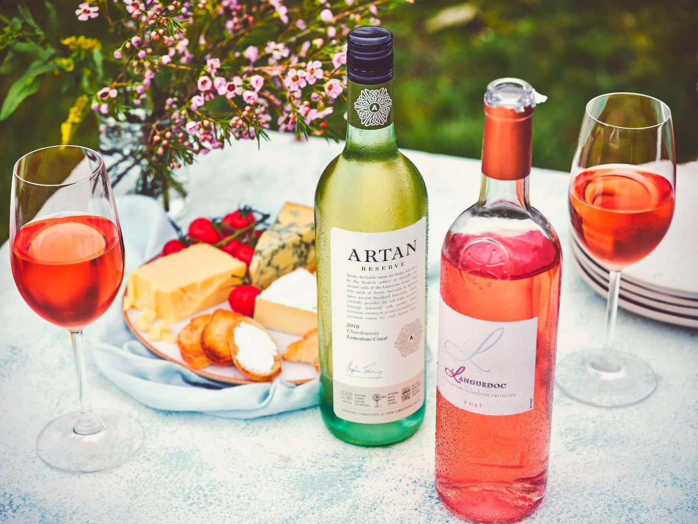 A picnic table set with a bottle of white wine, a bottle of rosé wine, two glasses of rosé, a cheese platter with assorted cheeses, grapes, and crackers, and a bouquet of pink flowers in the background.