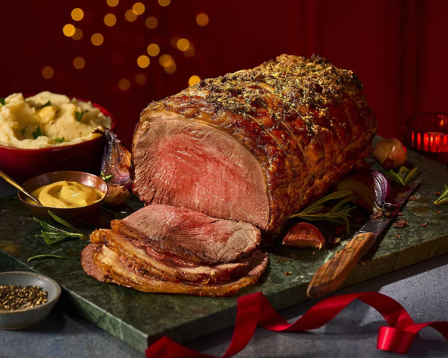 A plated roasted beef tenderloin with slices cut, served with mashed potatoes and mustard, garnished with herbs, on a dark surface with holiday lights in the background.