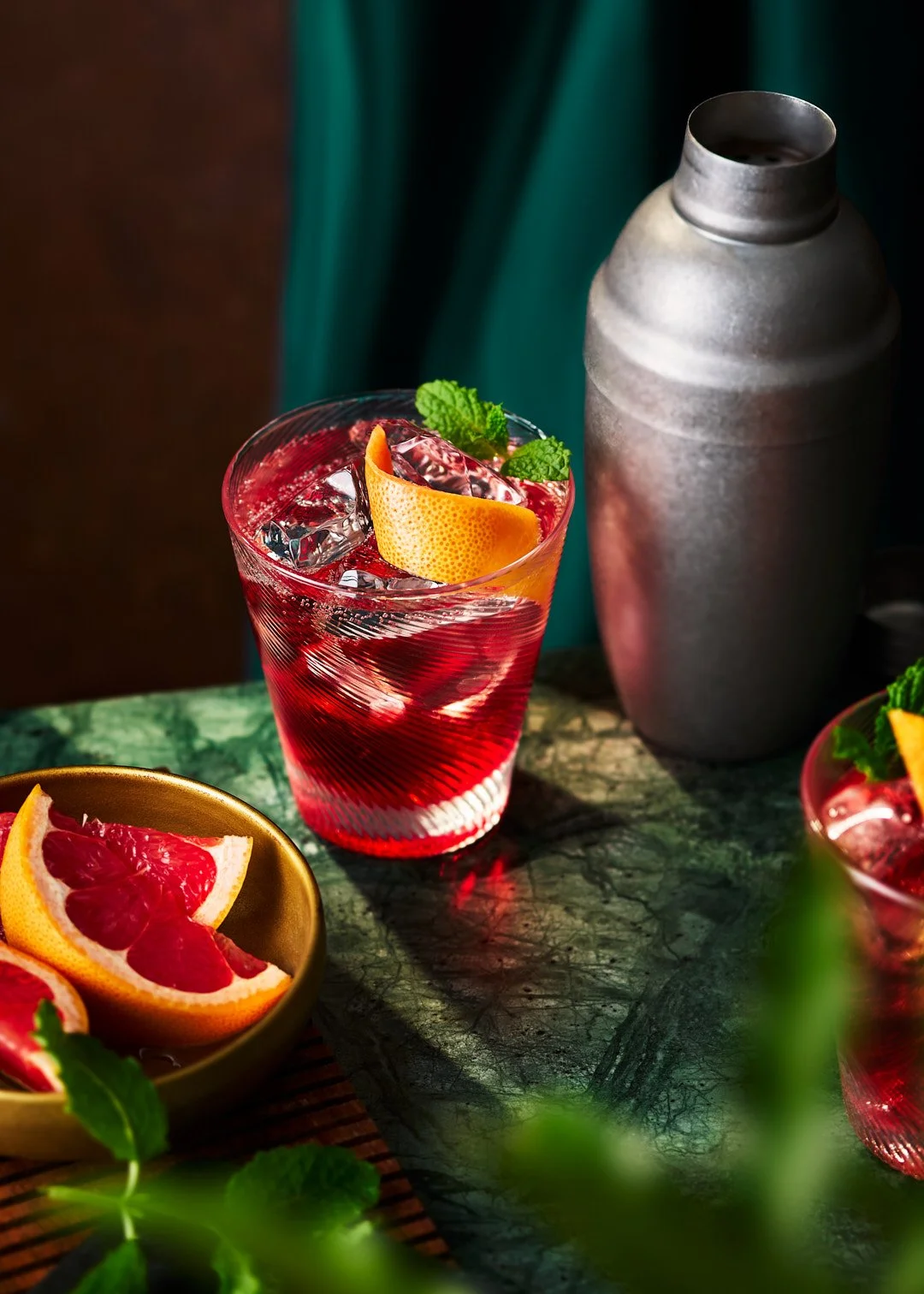 A glass of red cocktail with ice, garnished with an orange peel and mint, placed on a green marble surface. A bowl of grapefruit slices and a gray ceramic bottle are in the background.