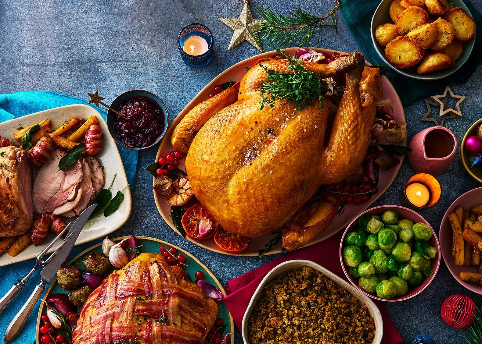 A festive feast with a roasted turkey, stuffing, roasted vegetables, green Brussels sprouts, roasted potatoes, cranberry sauce, and ham, decorated for the holidays with candles, ornaments, and greenery.