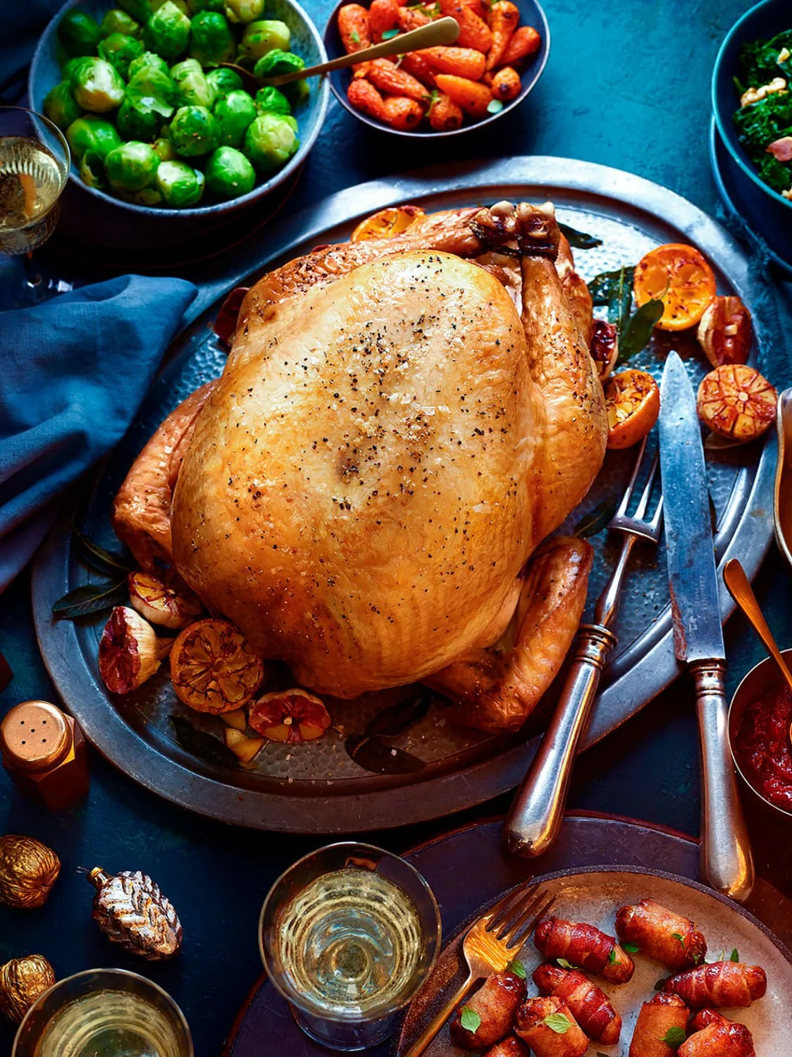 A roasted whole turkey with herbs and vegetables on a platter, surrounded by bowls of Brussels sprouts, carrots, and various side dishes, with glasses of white wine.