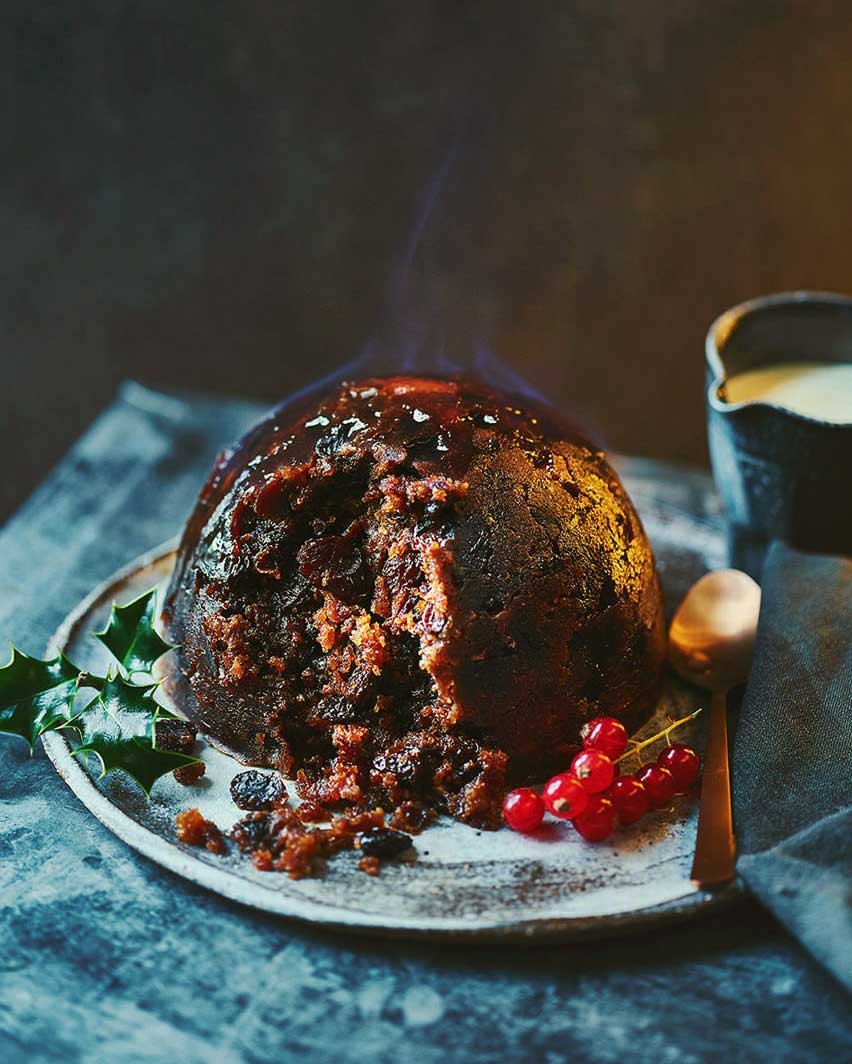 A steaming Christmas pudding with holly garnish on a white plate, accompanied by red currants, a spoon, and a glass of cream on a dark table.