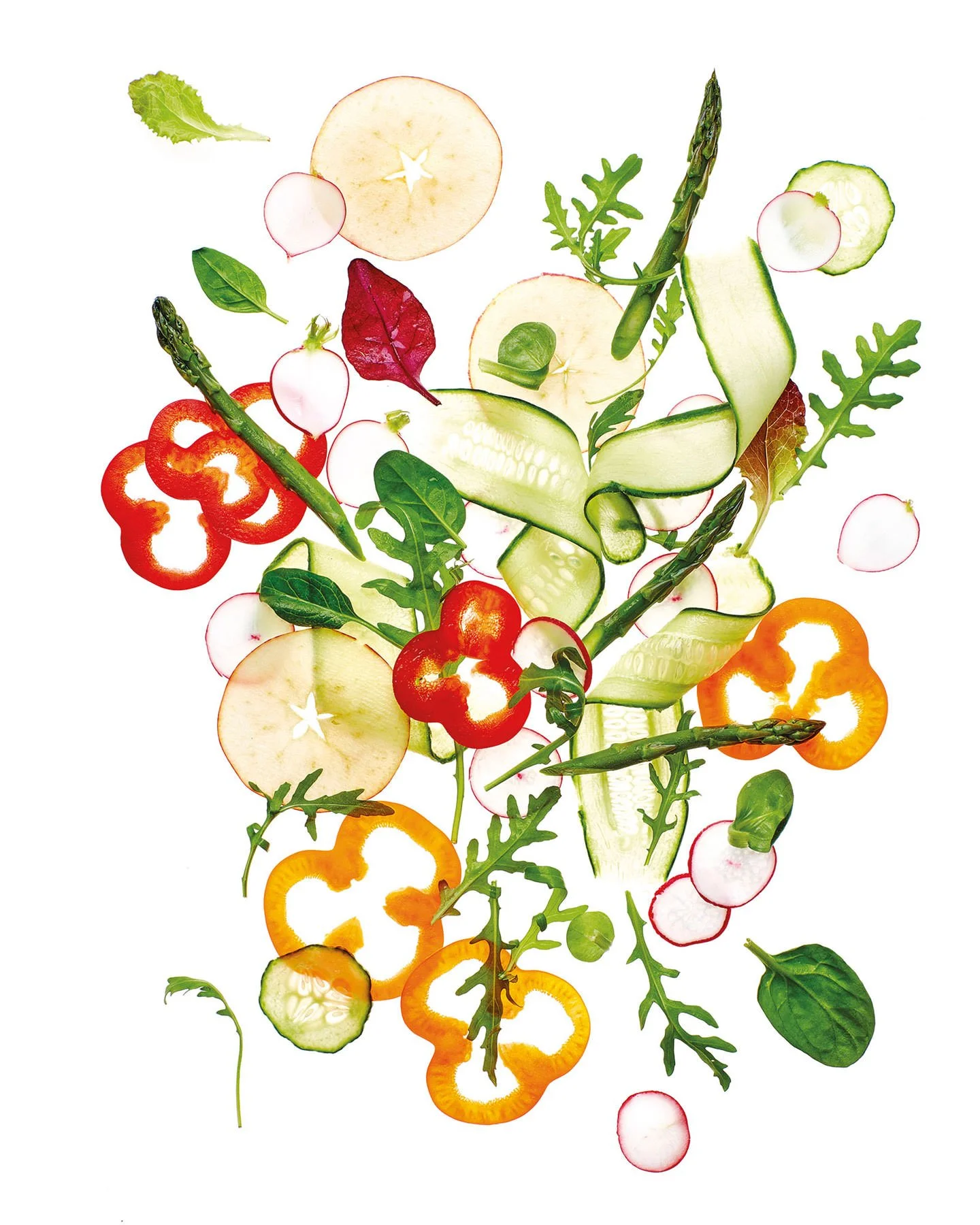 Sliced vegetables and greens including apples, radishes, cucumbers, bell peppers, asparagus, basil, arugula, and spinach on a white background.