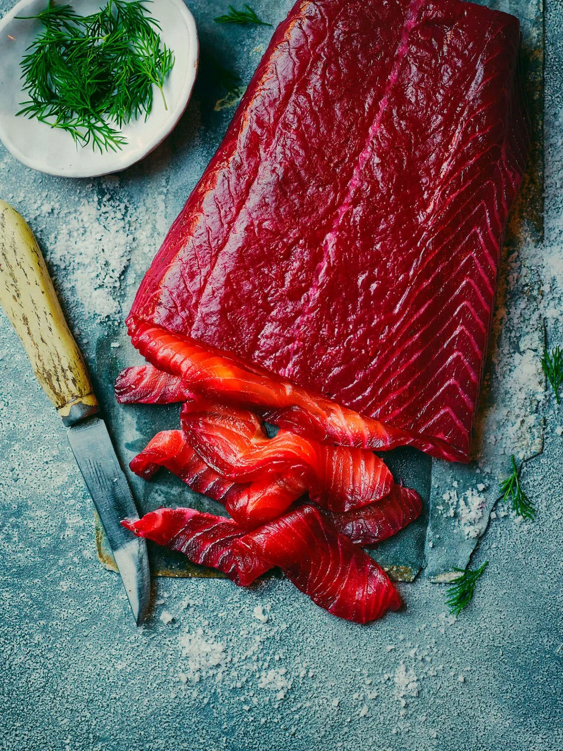Fresh raw red fish fillet on a dark stone surface with sliced fish pieces, a knife with a wooden handle, a small dish of green herbs, and scattered green sprigs.