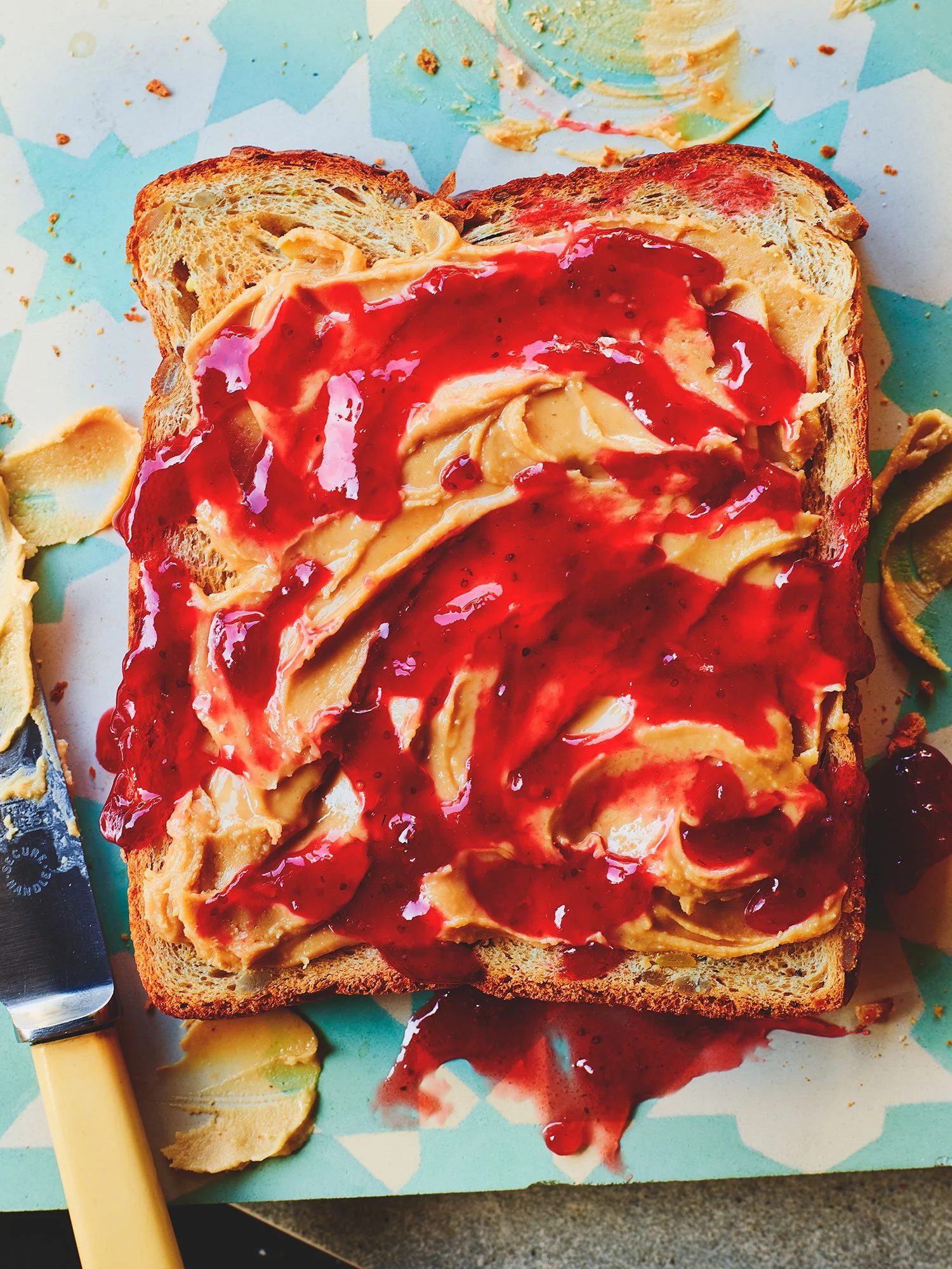 A slice of toasted bread with peanut butter and red jelly on top, served on a colorful paper with a butter knife beside it.
