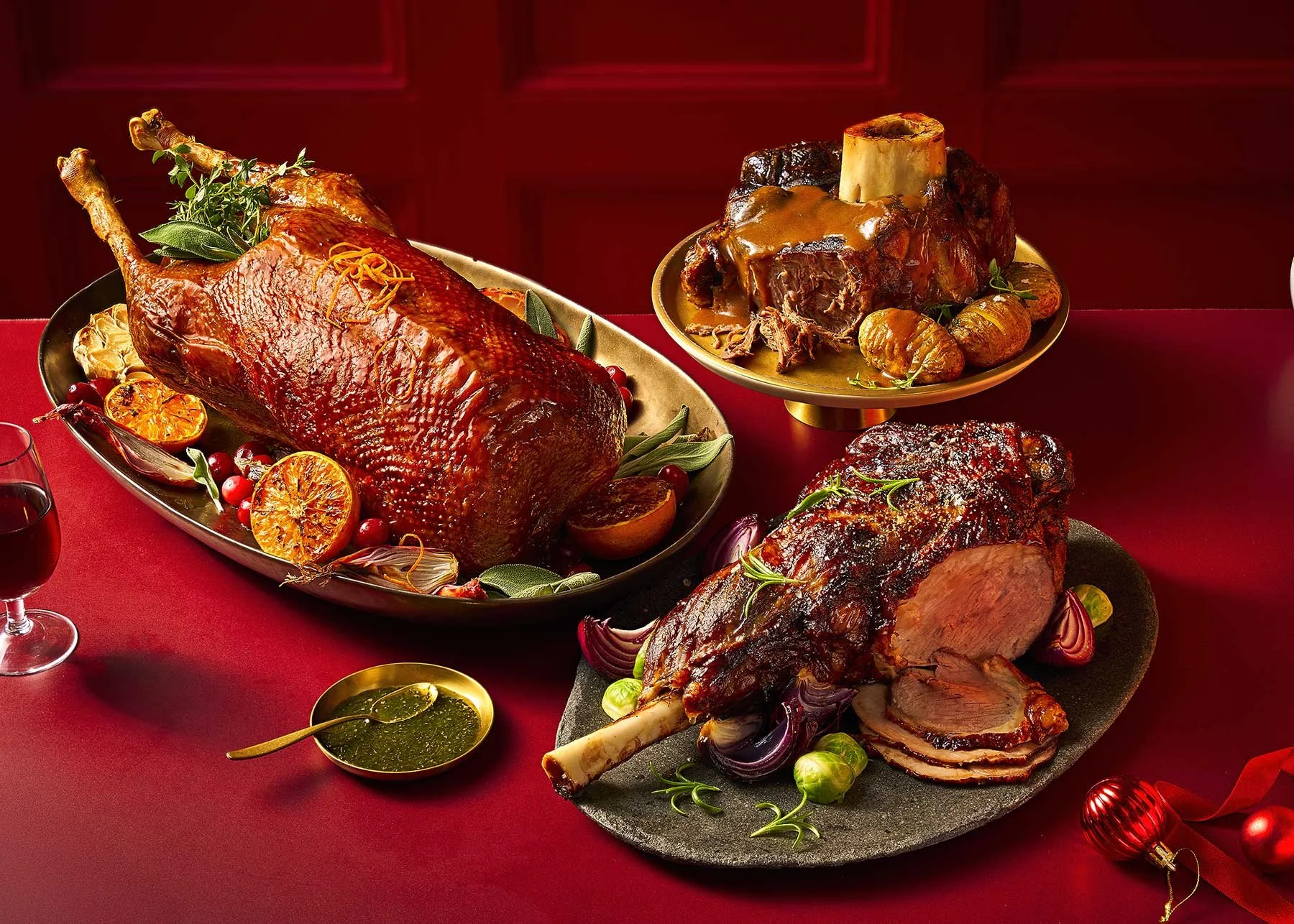 Festive table with roasted ham, lamb shank, and a meat dish with vegetables, set on a red tablecloth with wine and holiday decorations.