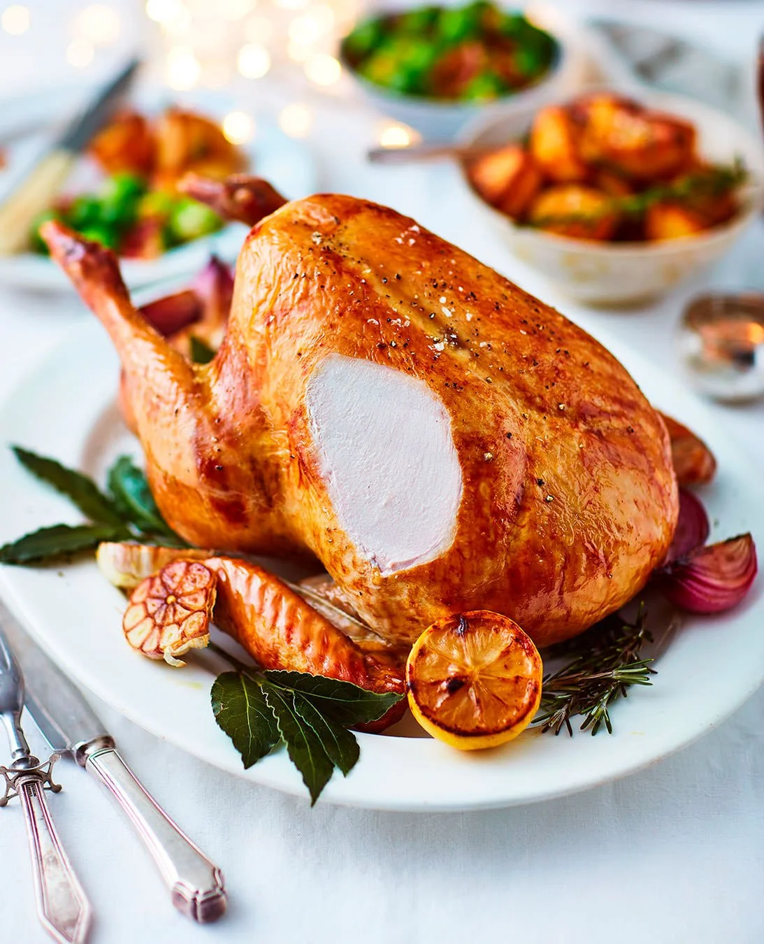 Roast chicken with golden, crispy skin garnished with herbs and lemon slices on a white platter, surrounded by dishes of vegetables and salads.