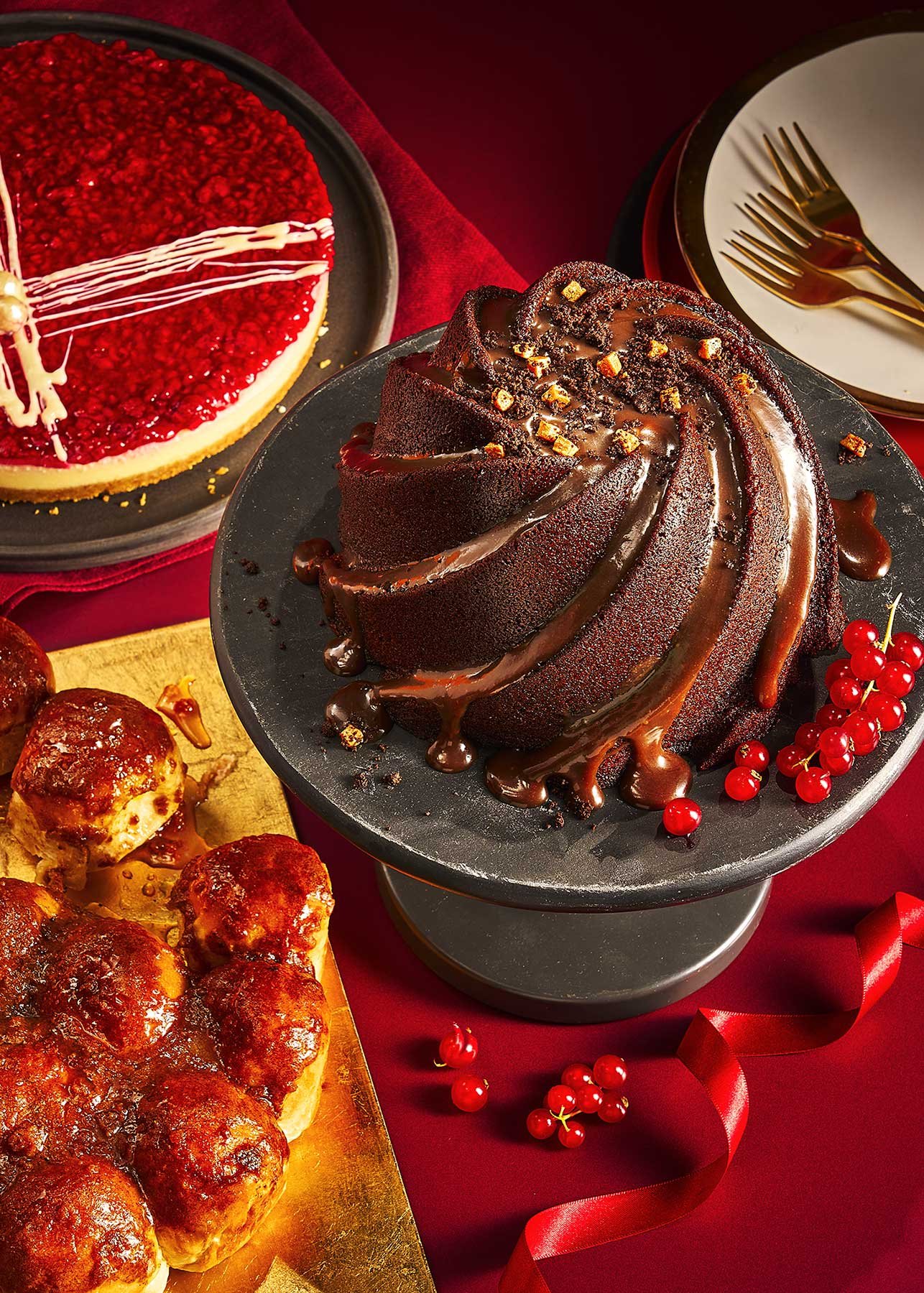 A table with a chocolate bundt cake drizzled with chocolate syrup and garnished with red berries, a red velvet cake with white and red decorations, and a tray of baked meatballs, set on red tablecloth with gold utensils and plates.