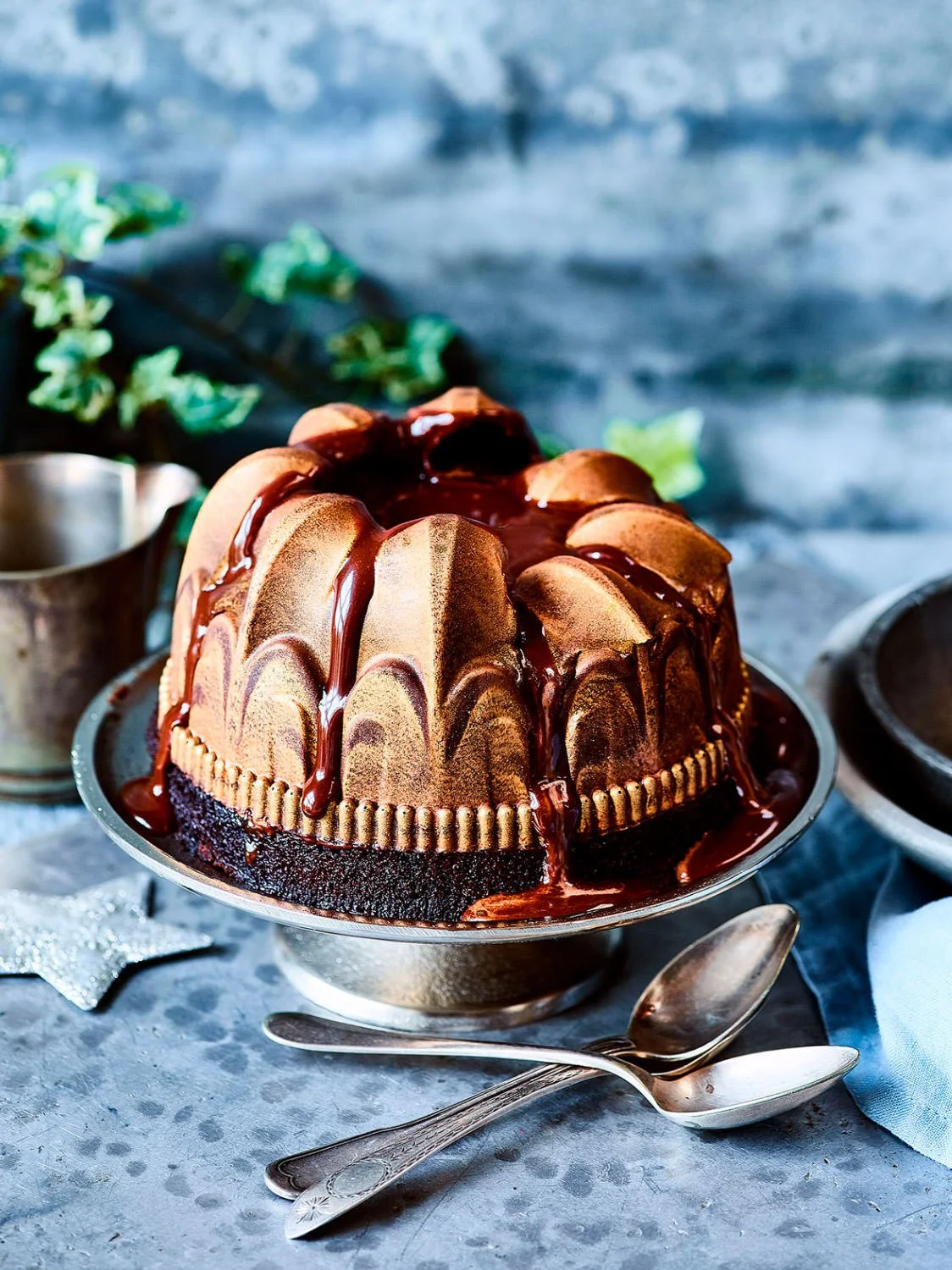 Chocolate bundt cake with glaze on top on a silver cake stand, with a silver spoon beside it, on a textured surface with a blue cloth and a green plant in the background.