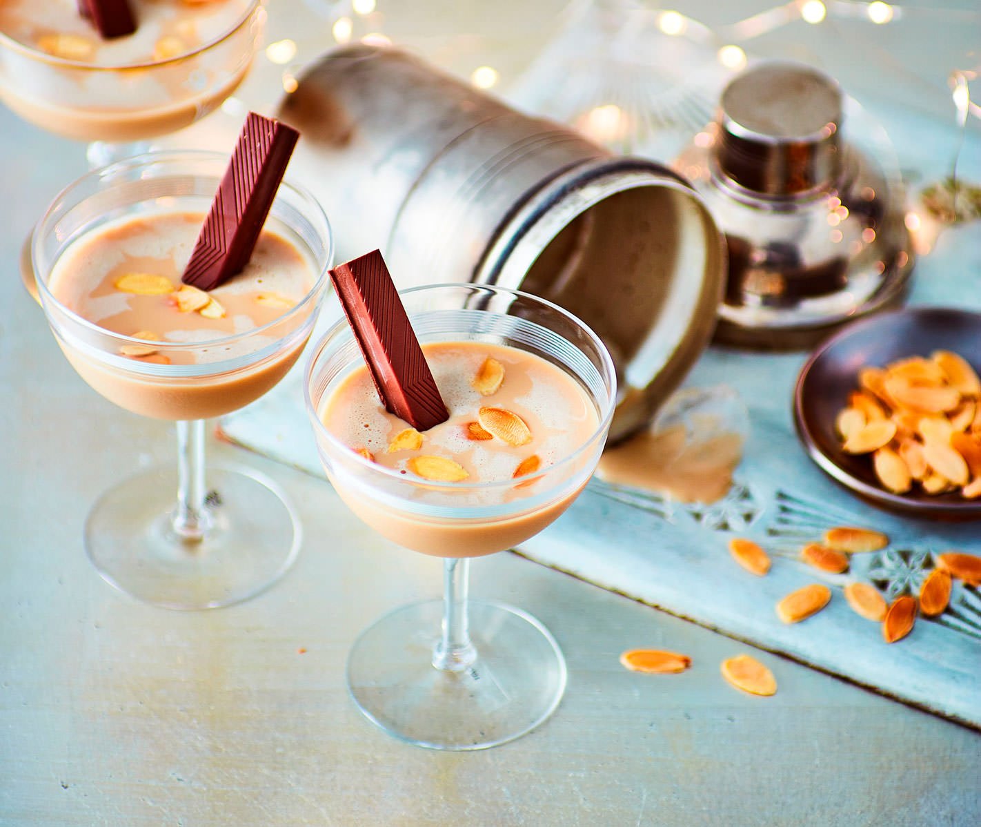Two glasses of pink beverage topped with almond slices and a piece of chocolate, set on a table with scattered almond slices and a silver container in the background.
