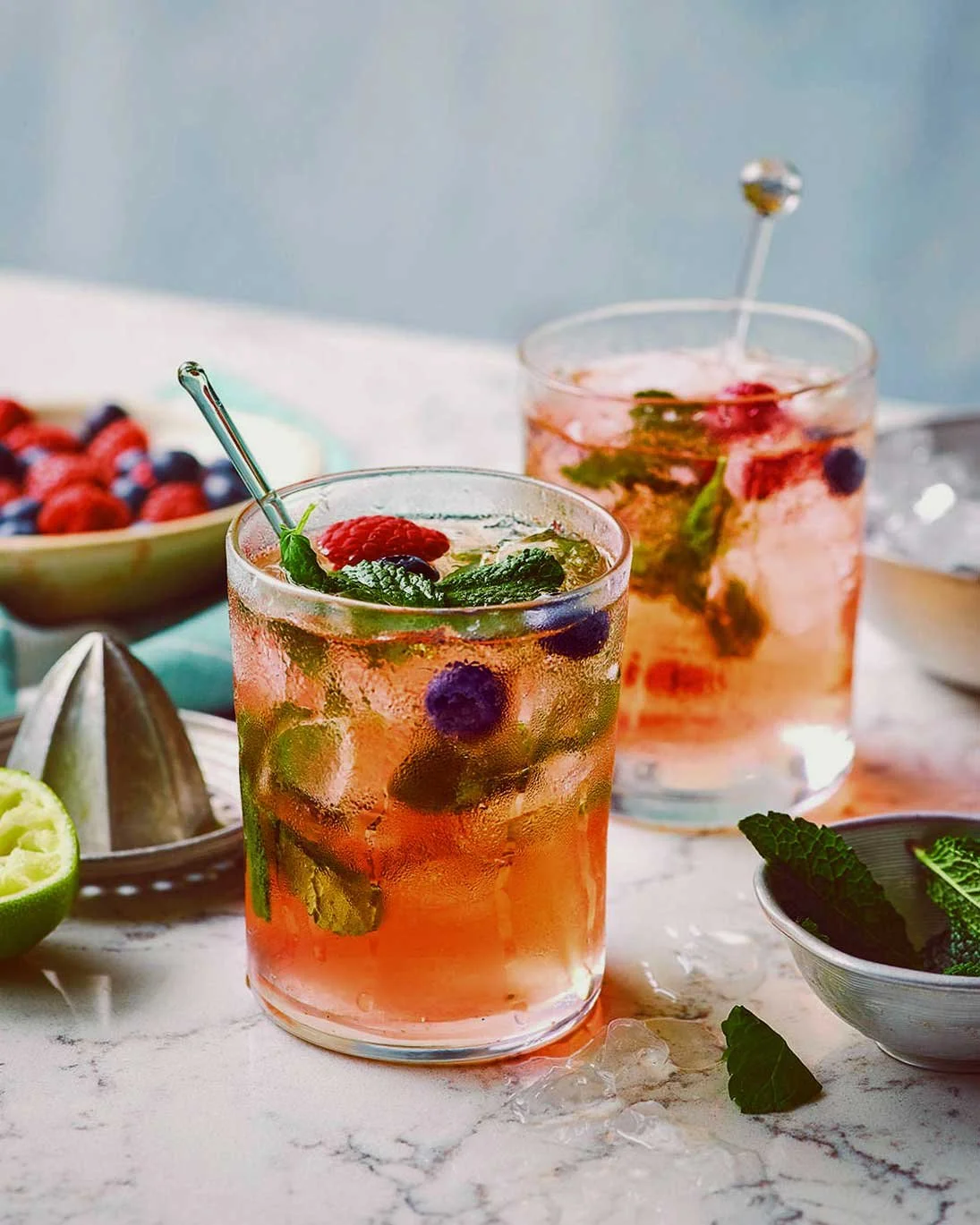 Two glasses of berry-infused beverages garnished with mint and berries, with bowls of berries, lime, and mint leaves in the background.