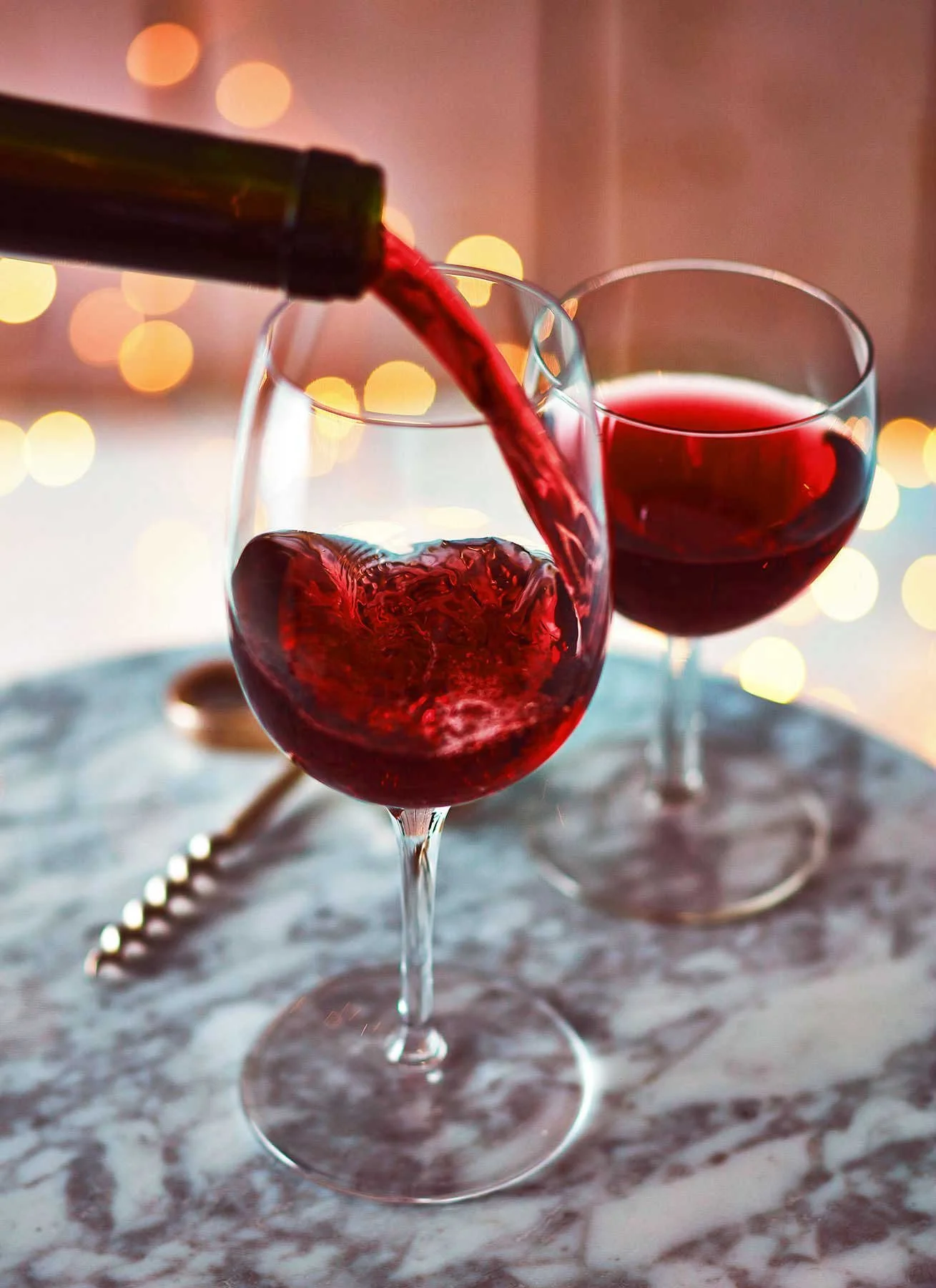 Red wine being poured into a glass, with another glass of red wine on a marble table, blurred warm lights in the background.