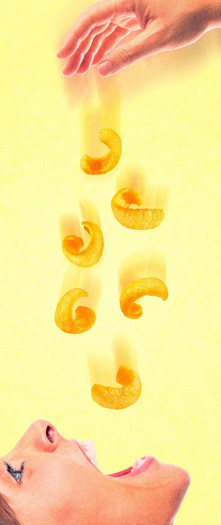 A woman with long hair and makeup is looking up and smiling at a hand dropping a snack curl into her open mouth against a yellow background.
