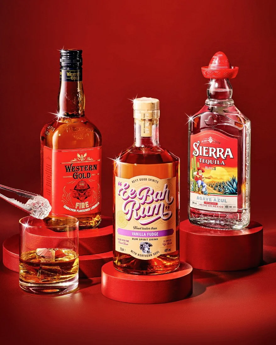 Three bottles of alcohol on red circular platforms, including Western Gold Fire cinnamon flavored liquor, E Bah Rum vanilla fudge rum, and Sierra Tequila blanco, with a glass of whiskey and a cocktail stick with sugar on the end.