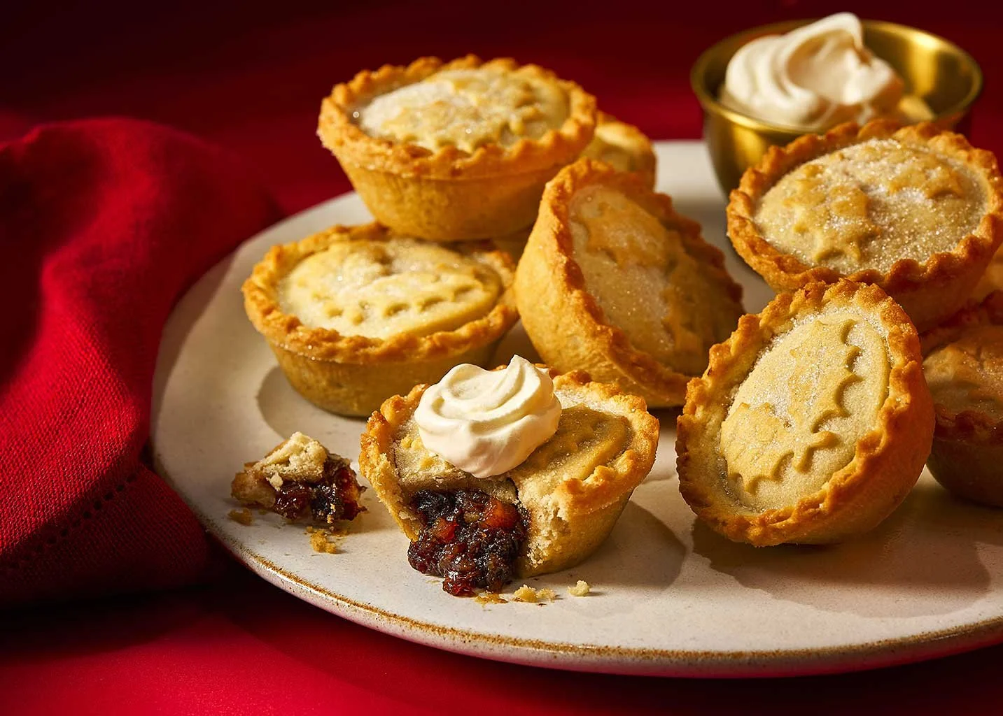 A plate of mini fruit pies with golden crusts, some topped with whipped cream and one with a piece taken out showing fruit filling, on a red cloth background.