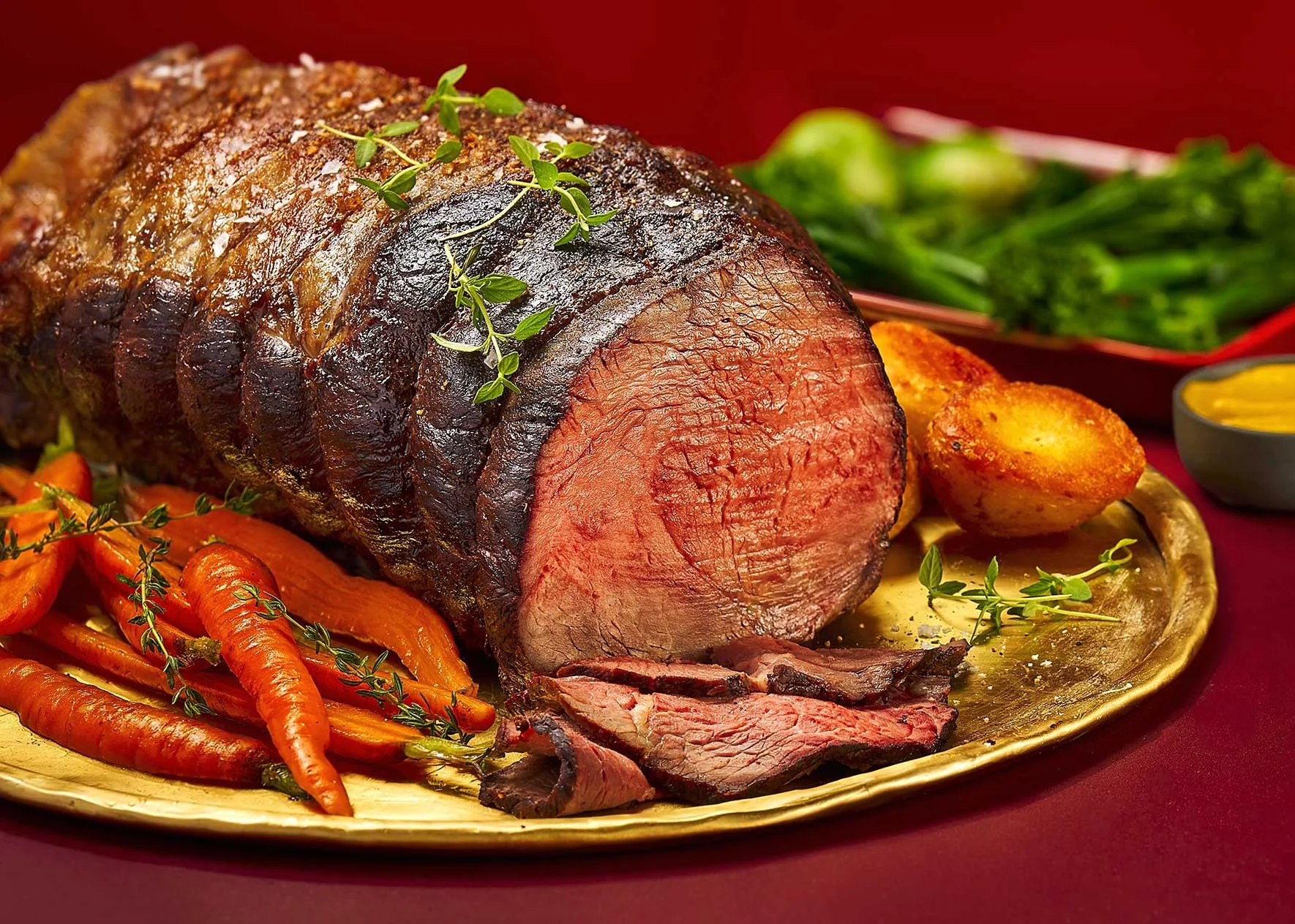 A roasted beef roast garnished with herbs, served with roasted carrots, a baked potato, green beans, and a small dish of mustard on a gold platter.