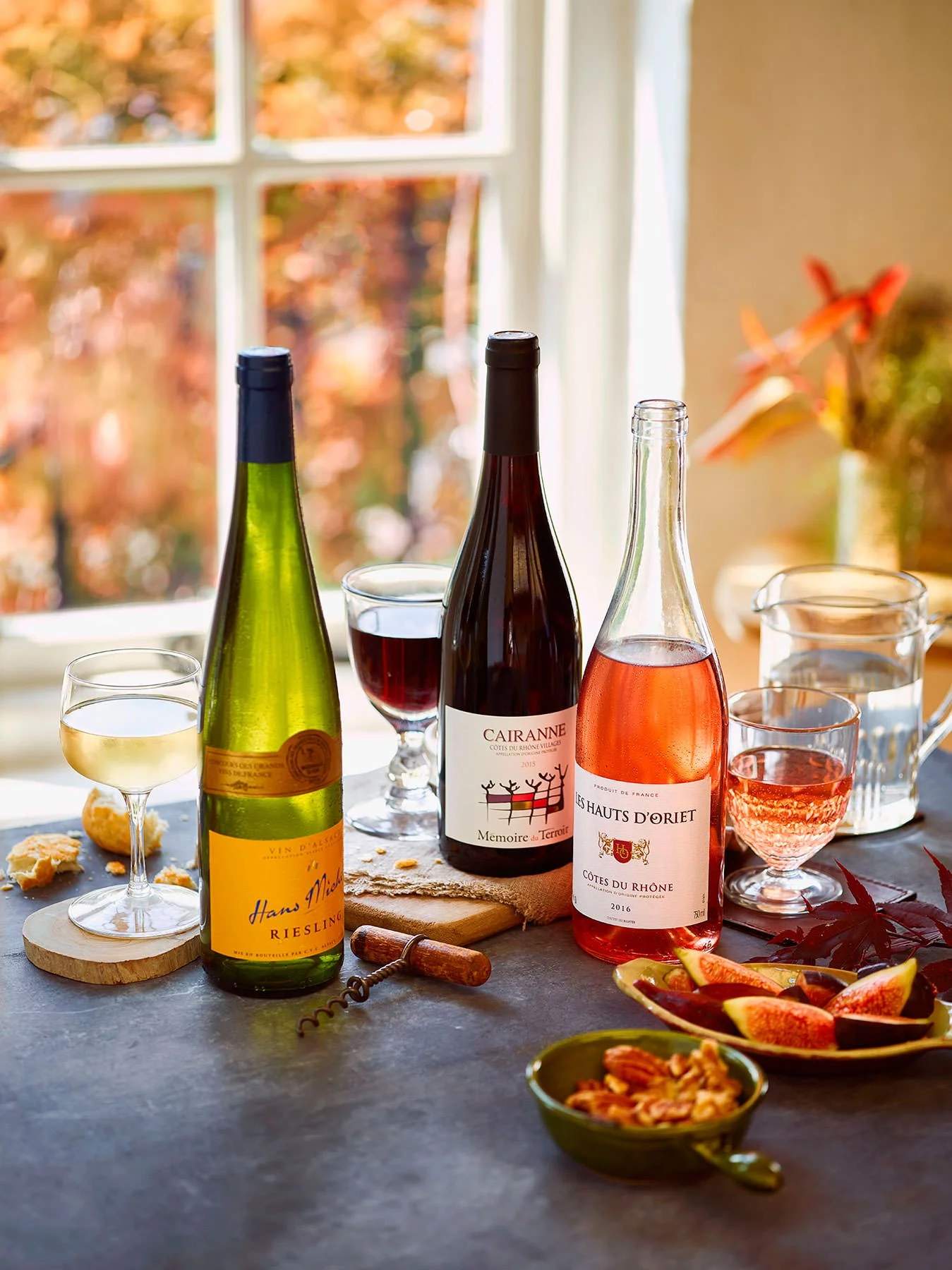 A table with three wine bottles, glasses of white, red, and rosé wine, a bowl of walnuts, and a plate of sliced figs, set near a window with a view of autumn foliage.