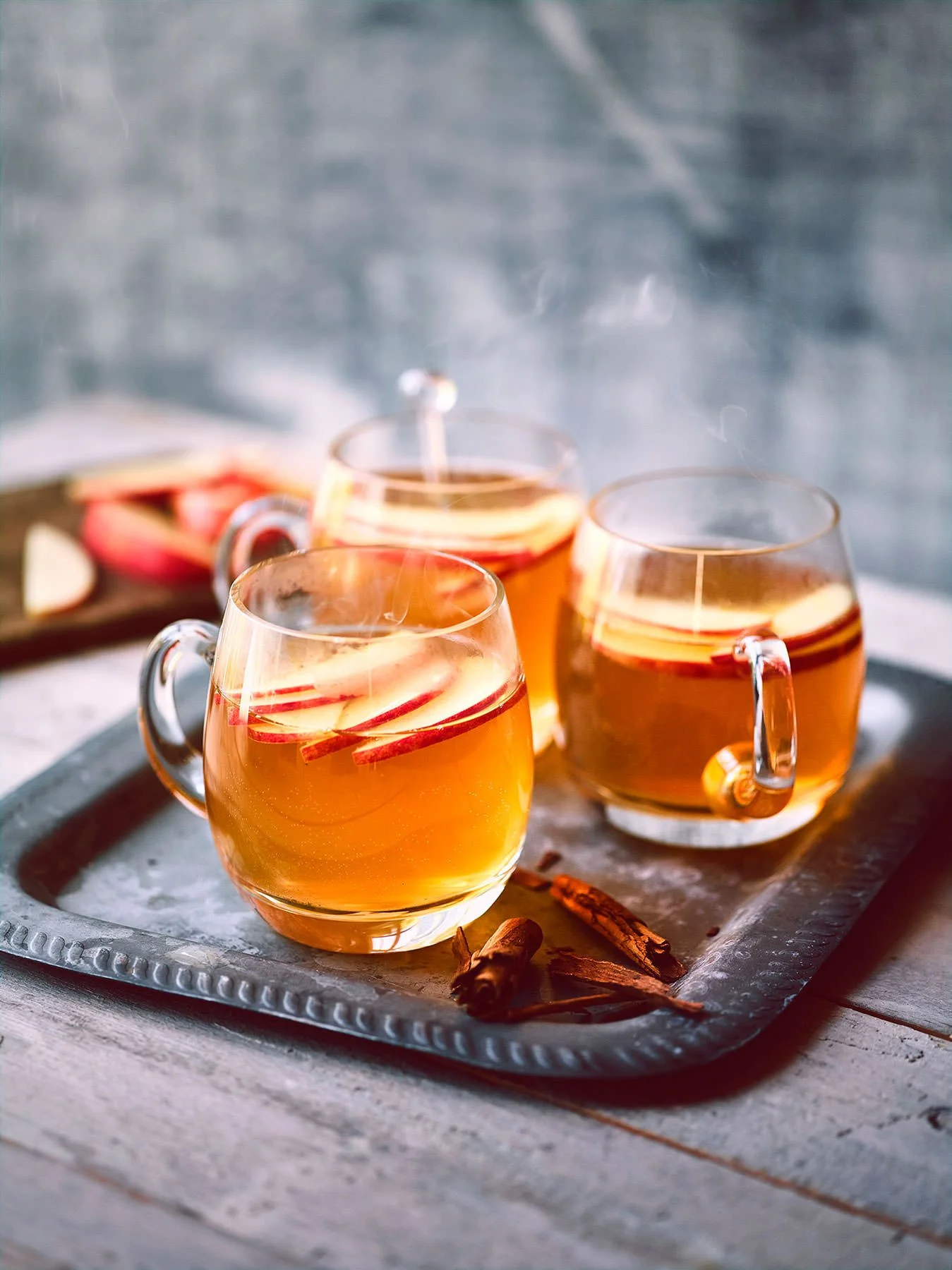 Three clear glass mugs filled with apple cider garnished with apple slices, placed on a silver tray with cinnamon sticks, on a rustic wooden surface.