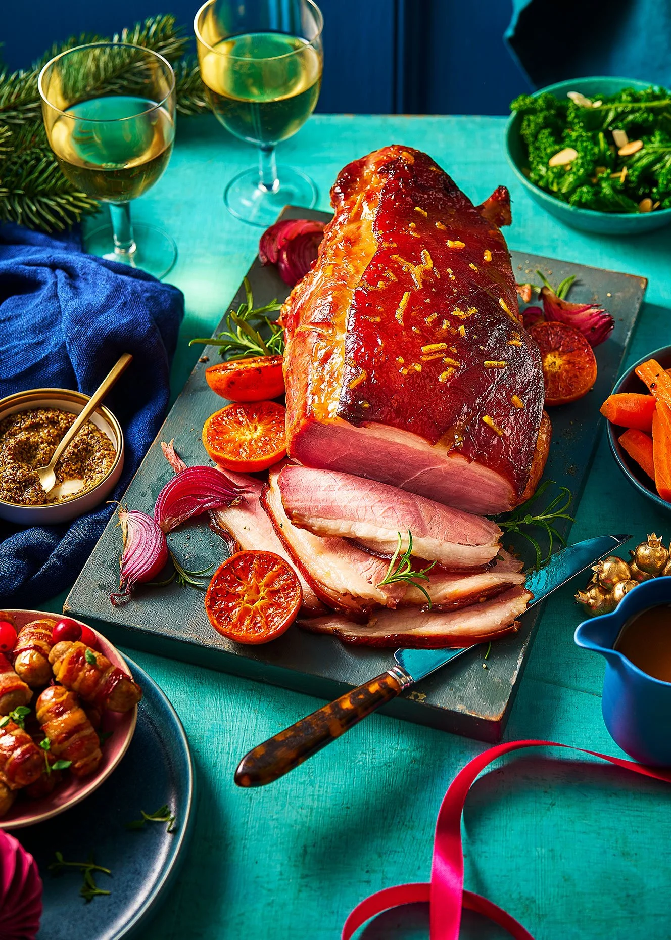Sliced glazed ham with pineapple and cherry garnishes, surrounded by sides including green salad and roasted vegetables, on a festive teal table with wine glasses.