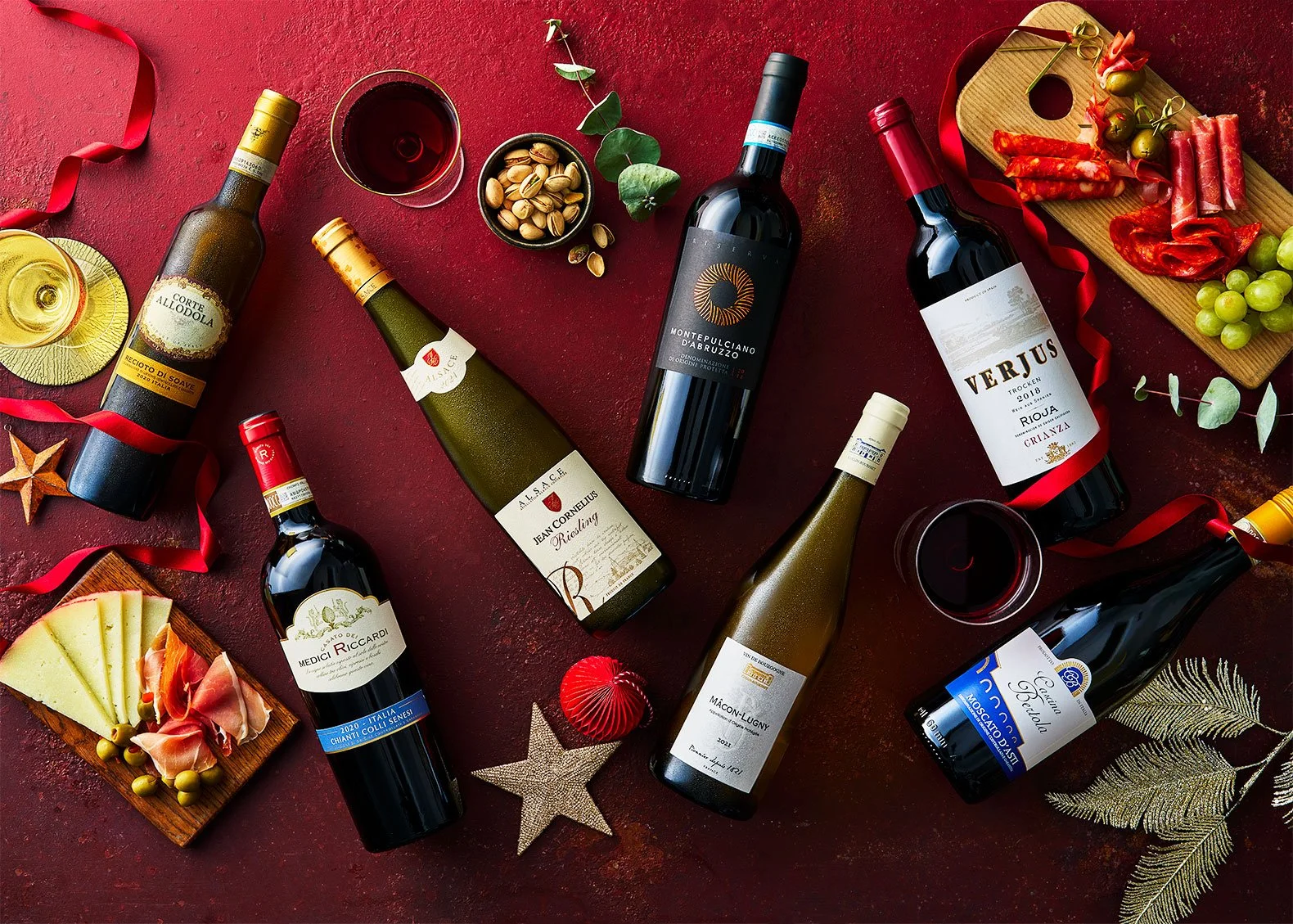 Multiple bottles of red wine, glasses filled with red wine, cheese, cured meats, grapes, pistachios, green leaves, and holiday decorations on a dark red background.